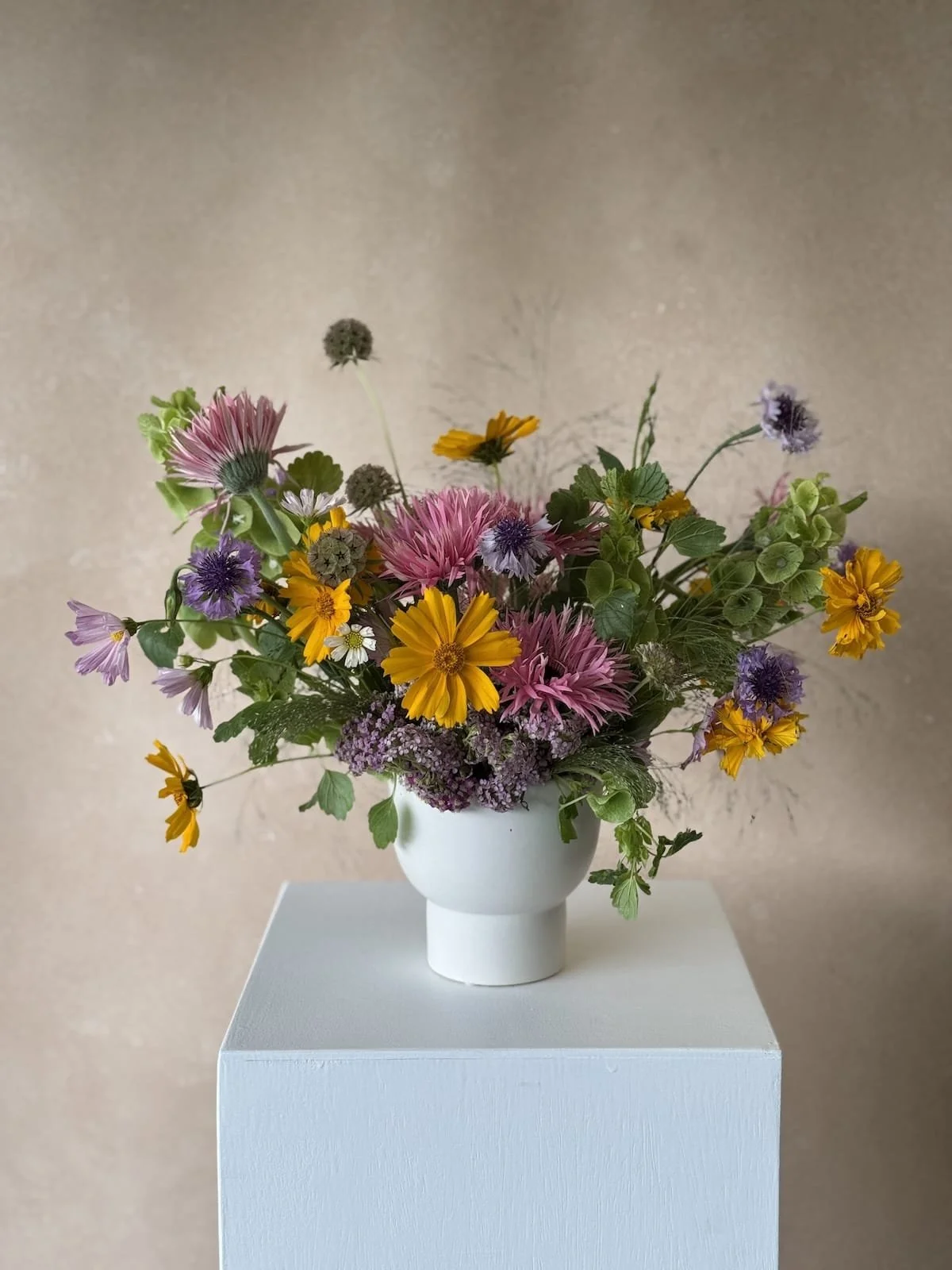 Colorful bouquet of pink, purple, yellow, and white flowers in a white ceramic vase on a white pedestal against a beige background.
