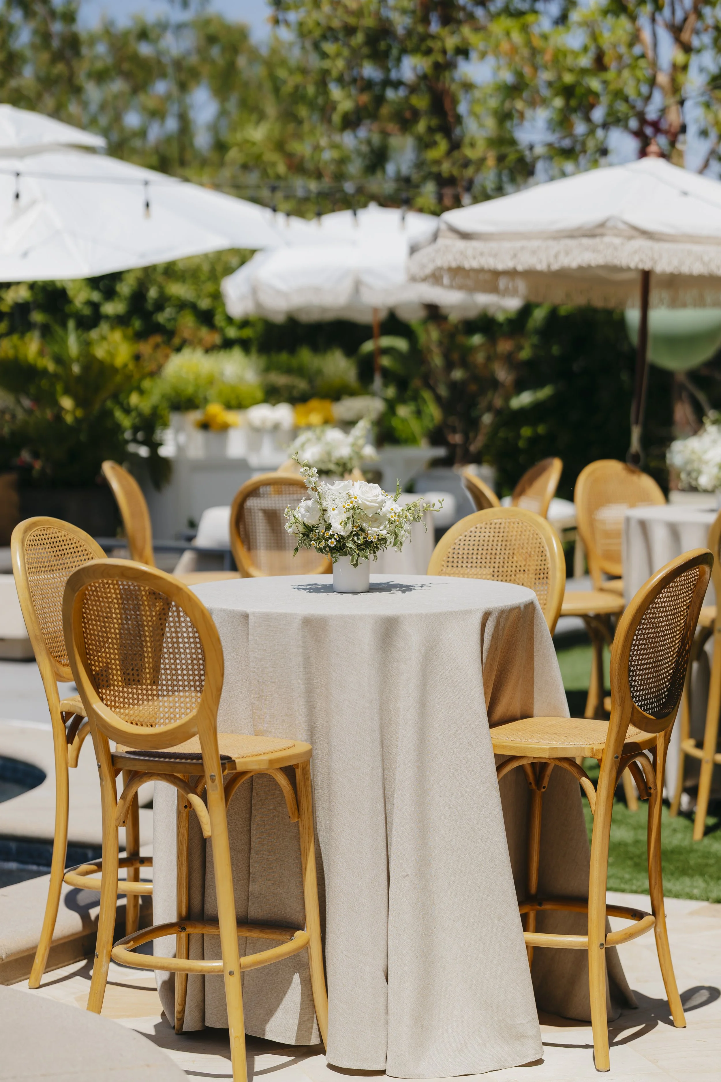 Outdoor dining setup with a round table covered with a beige tablecloth, a floral centerpiece, surrounded by rattan chairs, with white umbrellas and lush greenery in the background.