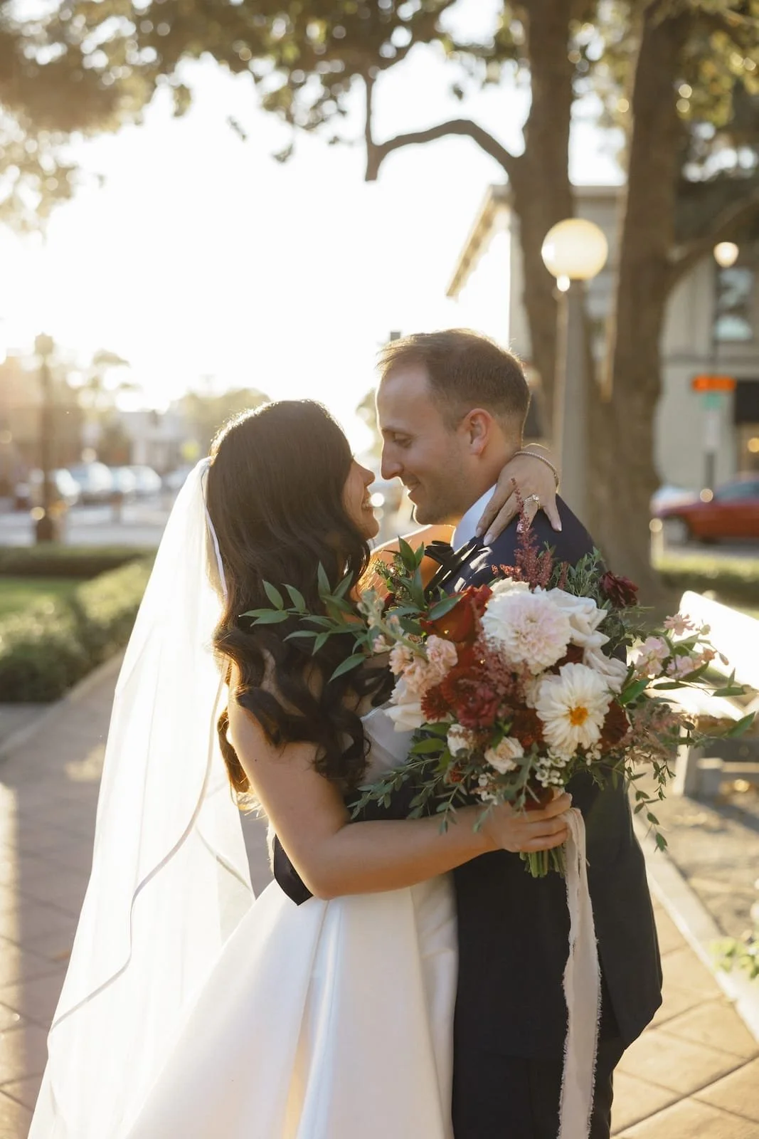 A bride and groom share a romantic moment outdoors during sunset; the bride holds a large bouquet of flowers, and they are close with their foreheads touching.