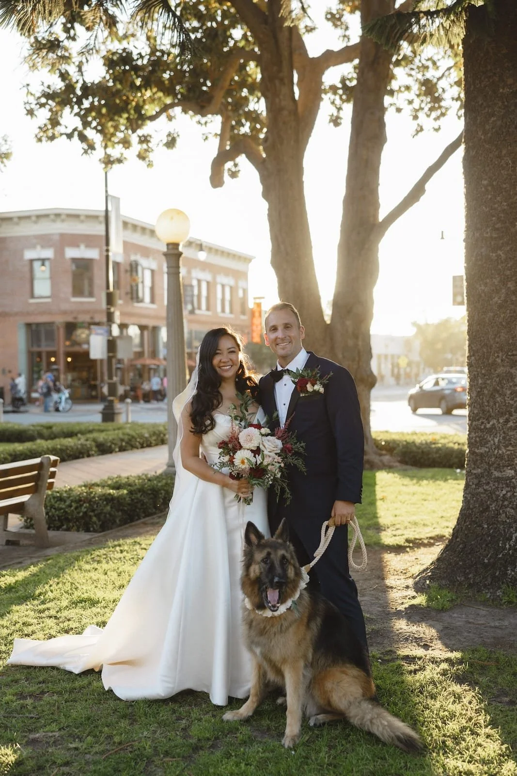 A bride and groom in wedding attire standing outdoors with a German Shepherd dog, holding a bouquet of flowers, under large trees with sunlight in the background.
