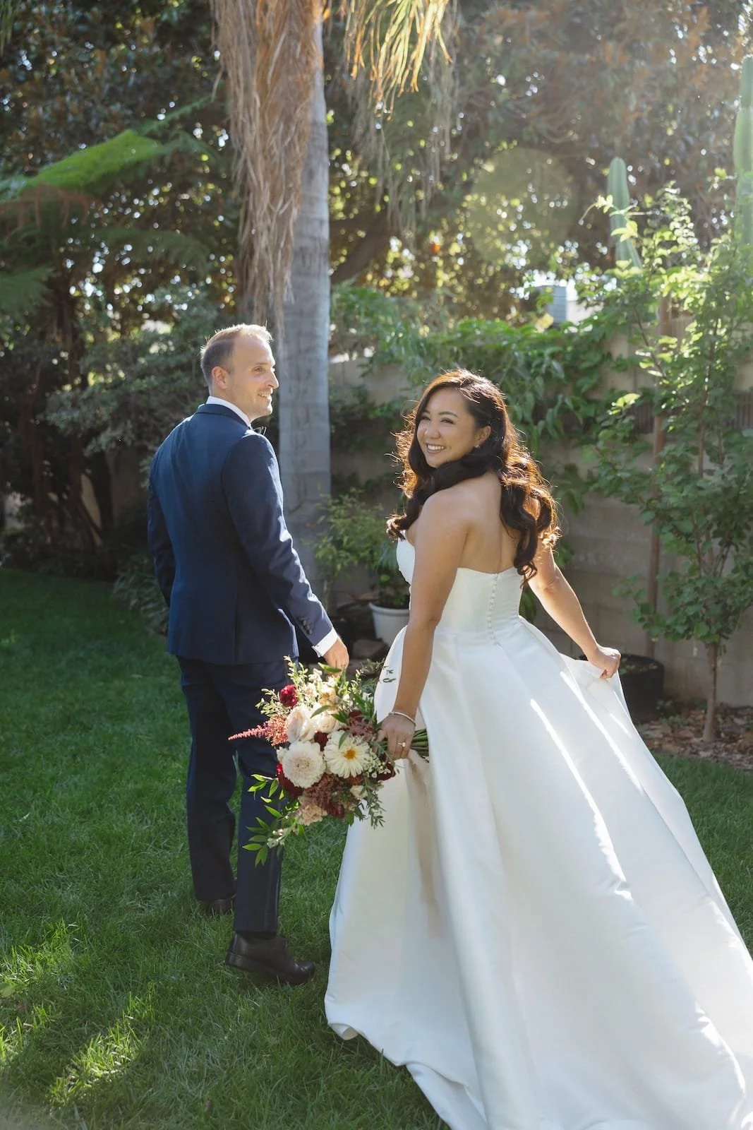 A bride and groom in a garden, the bride holding a bouquet and smiling at the camera, the groom looking back at her, with sunlight filtering through trees.