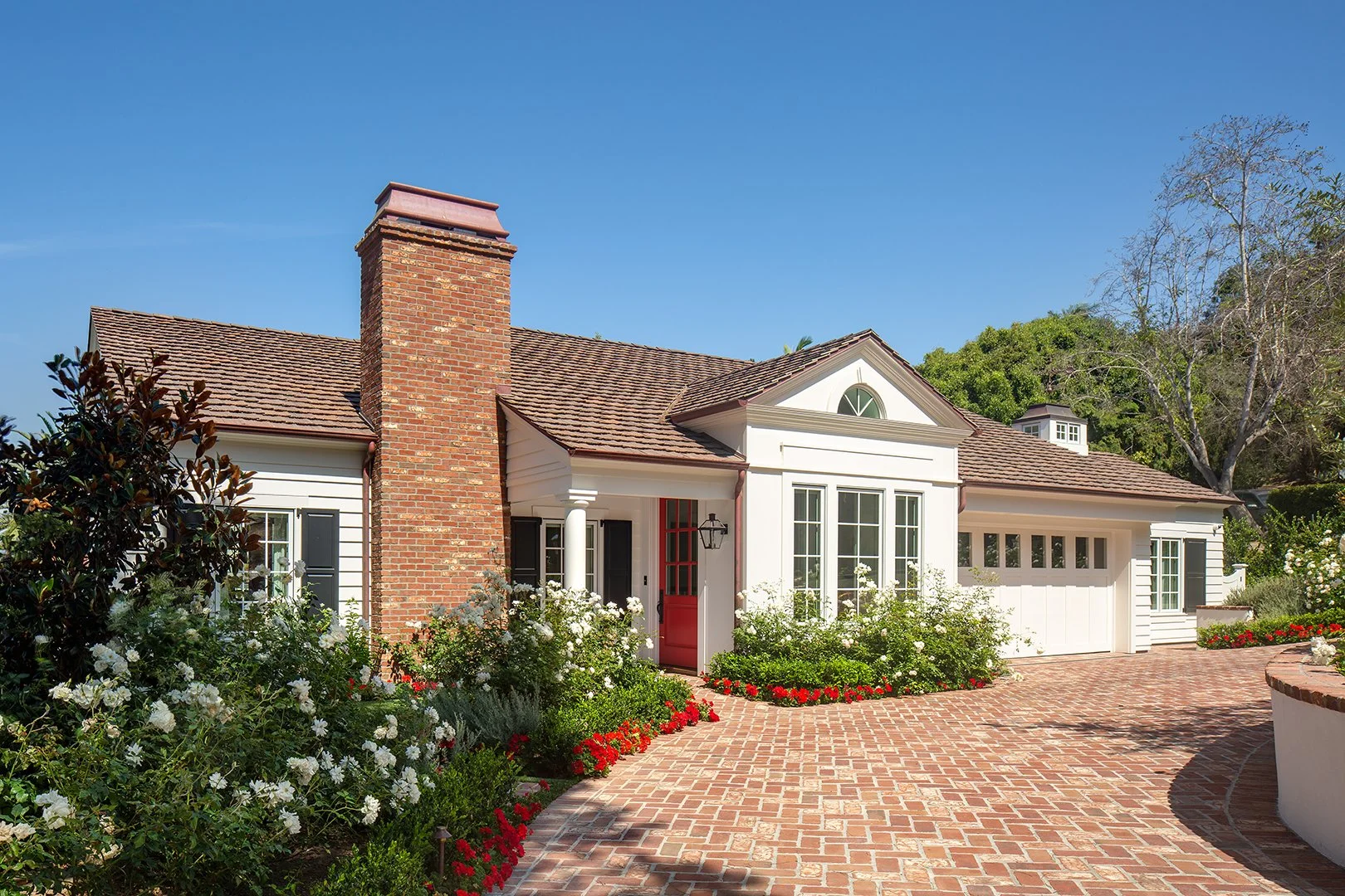 Front view of a house with a brick driveway, white exterior, red front door, brick chimney, and lush green plants and flowers.