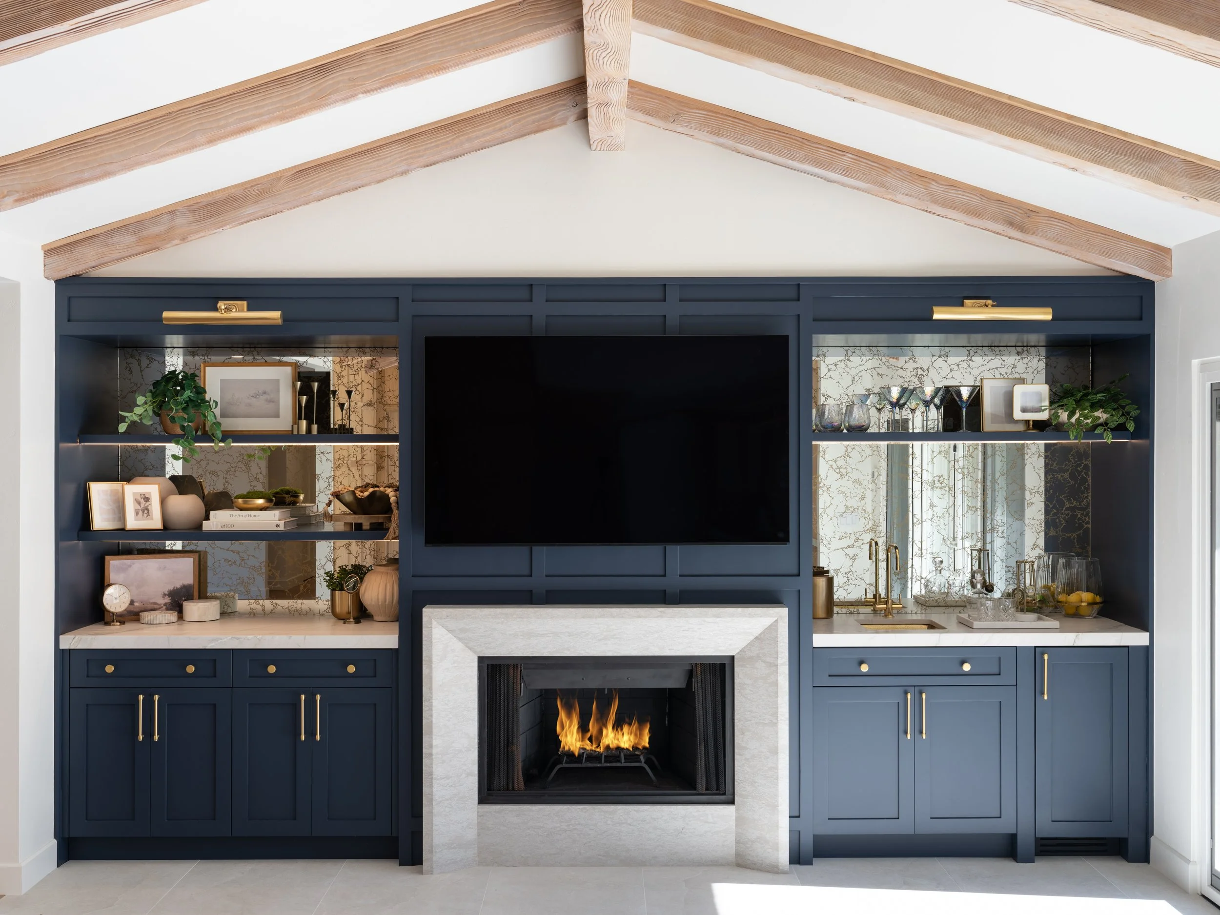 Living room with navy blue built-in shelving unit, television, and a fireplace with a burning fire, against a wall with patterned wallpaper and wooden ceiling beams.