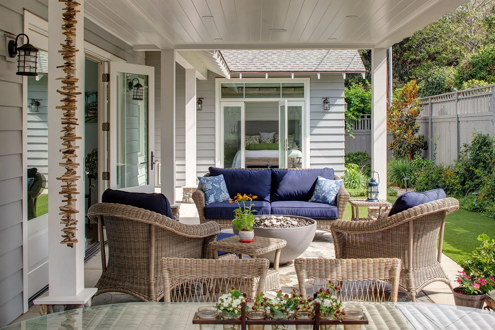 Covered outdoor patio with wicker furniture, navy cushions, potted plants, and garden view.