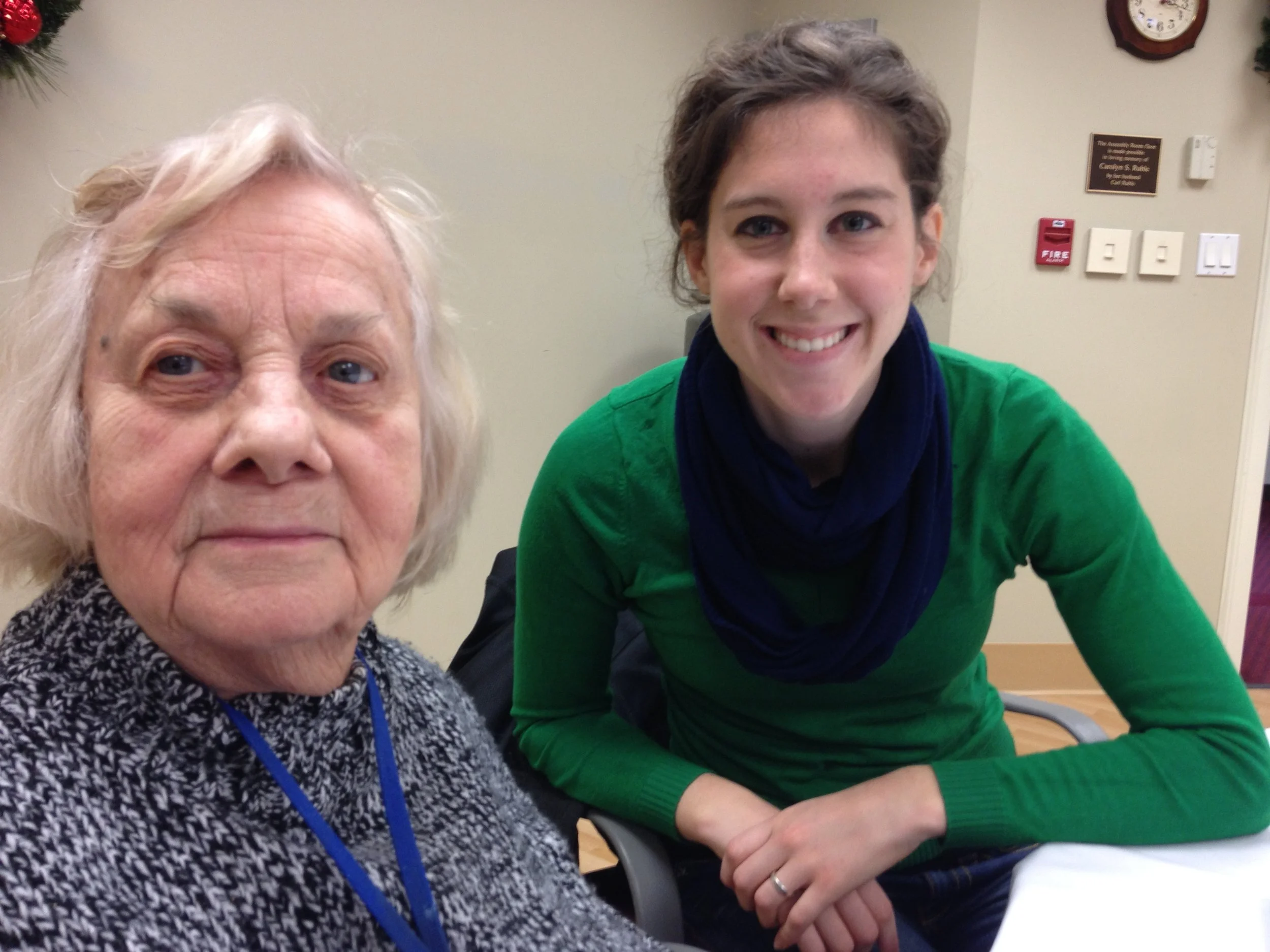An elderly woman with gray hair and a patterned sweater sitting next to a young woman with short brown hair, wearing a green top and blue scarf, smiling at the camera in an indoor setting.