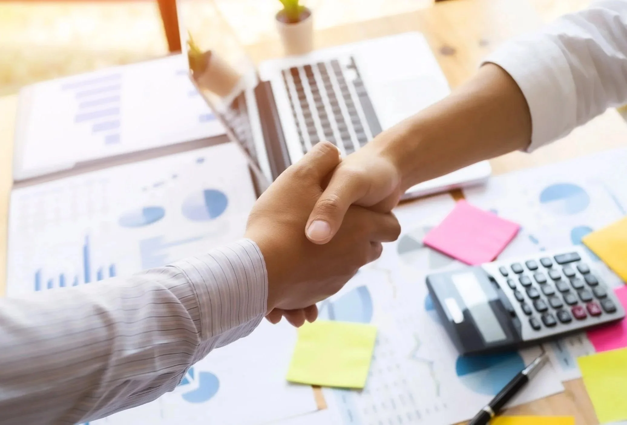 Two people holding hands over a desk with charts, a calculator, sticky notes, and a pen, in an office setting.