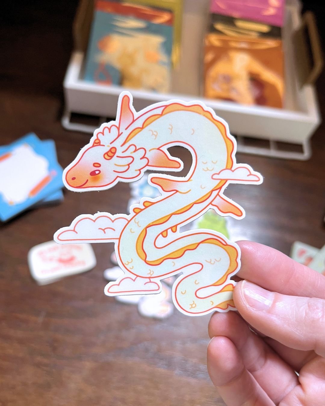 A hand holding a sticker of a cute cartoon dragon with a long, wavy body, a smiling face, and a small wing, surrounded by clouds.