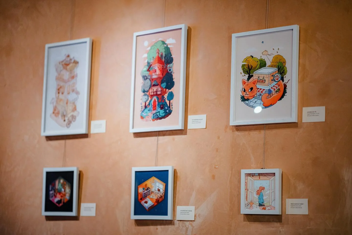 A display of seven framed illustrations on a wall, showcasing colorful animated art with various scenes, including a house, a tower, a cat with trees, an interior room, and a girl looking out a window.