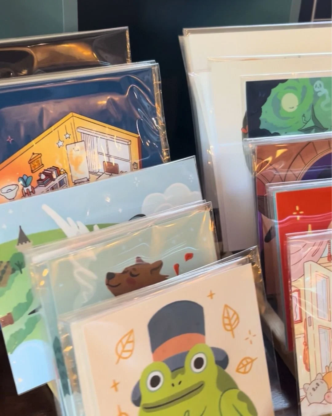 A display of various illustrated cards in plastic sleeves, featuring whimsical and colorful designs, including a frog wearing a top hat, a cozy room interior, a landscape with a castle, a bear, and a night sky with swirling green clouds.