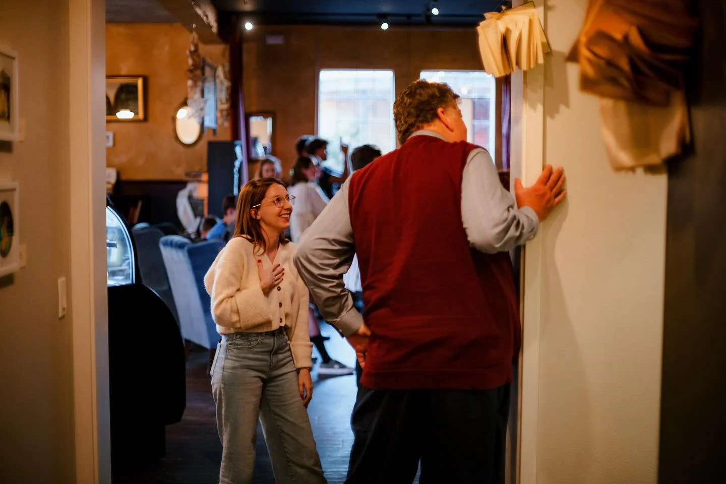 A young woman with glasses, wearing a beige sweater and gray pants, is smiling and placing her hand on her chest while talking to an older man. The man, seen from behind, is wearing a red vest and gray shirt. They are inside a warmly lit, cozy restau