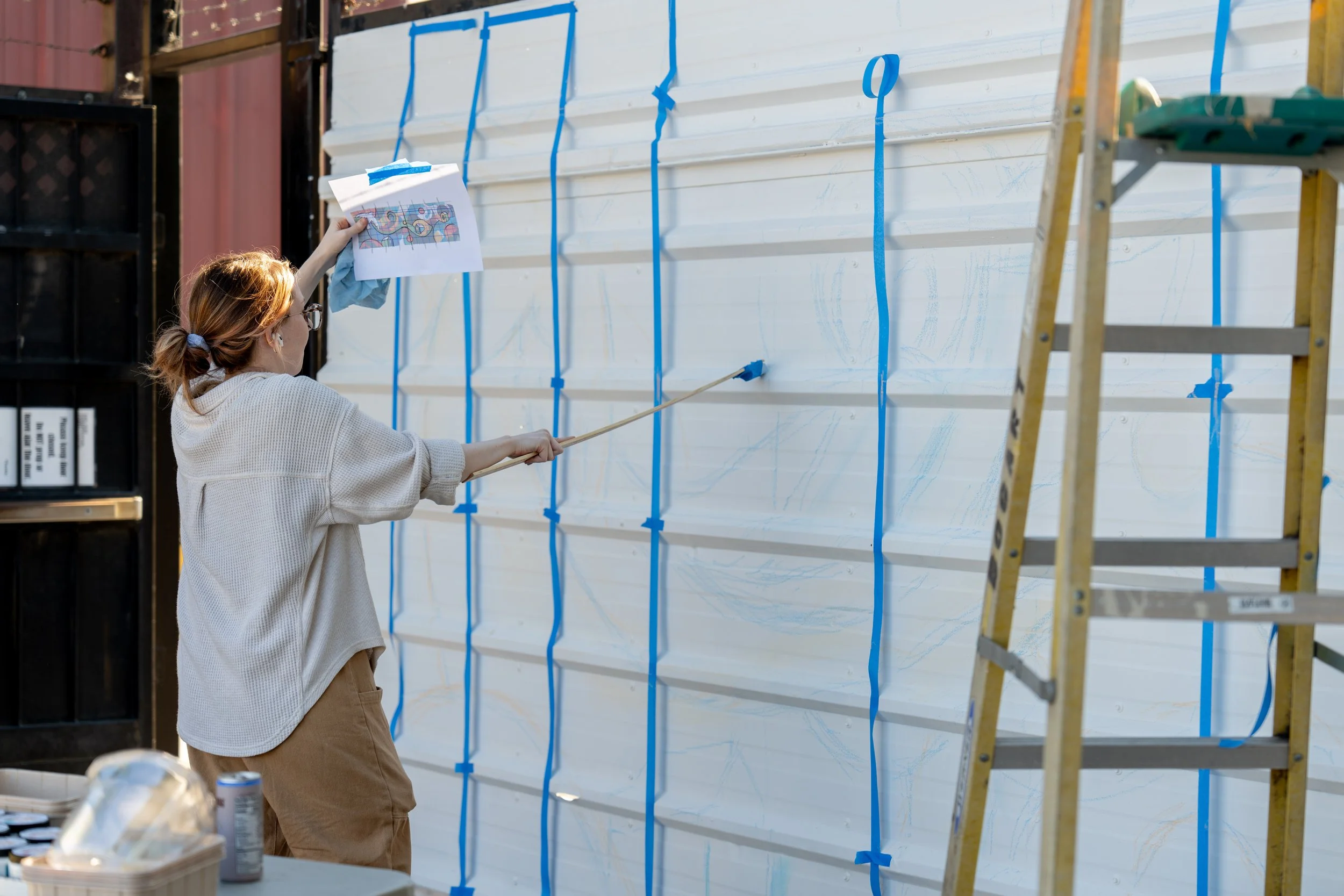 A woman painting a mural on a large white wall outdoors, holding a paintbrush and lifting a paper with a design, with a yellow ladder nearby.