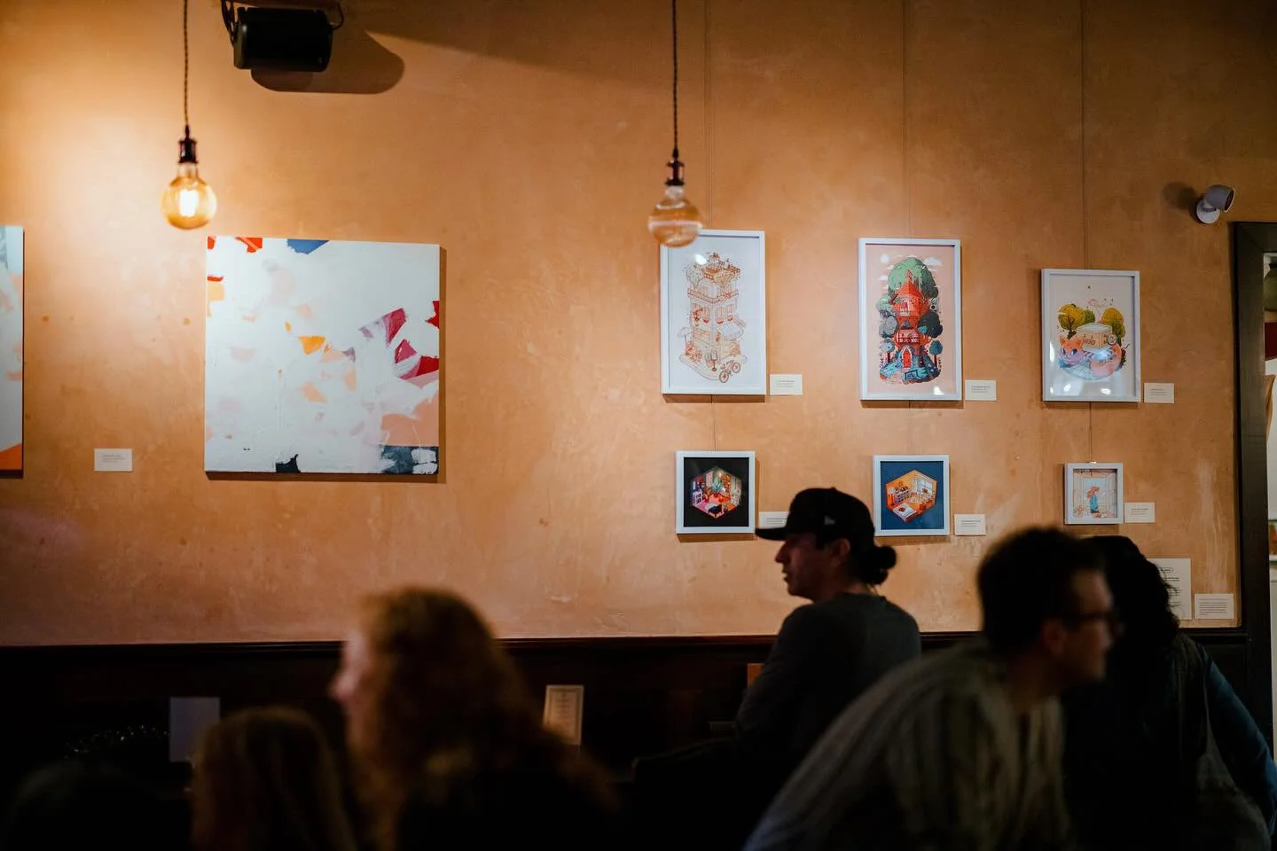 Art gallery wall with various framed illustrations of buildings and scenes, with a few people seated and chatting in the foreground.