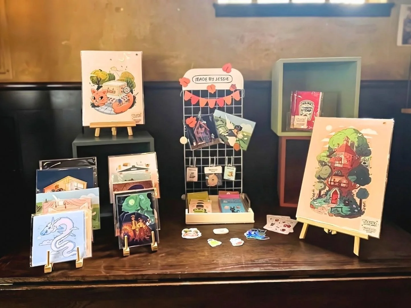 Display of greeting cards, stickers, and small artwork on a table with decorative stands and a wire rack, featuring colorful illustrations including animals, nature scenes, and imaginative designs.