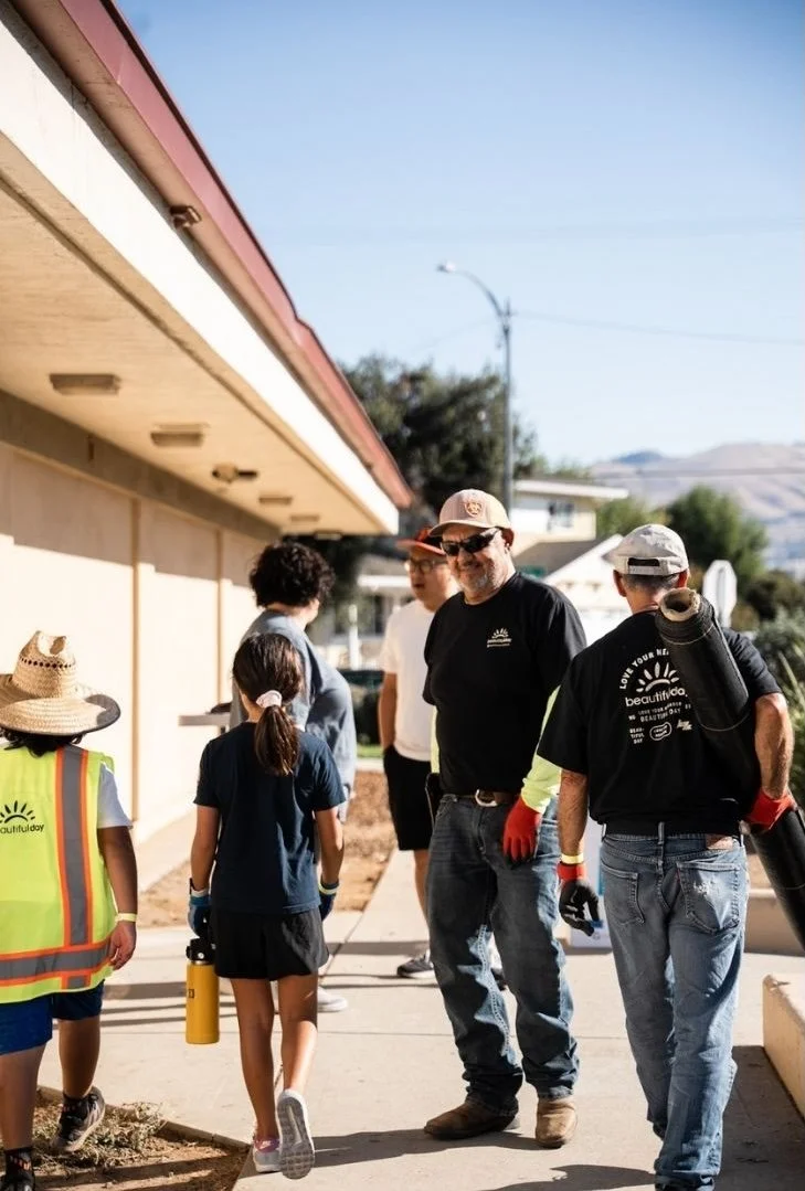 A group of volunteers helping clean up a sidewalk outside a building, wearing gloves and safety vests, on a sunny day.