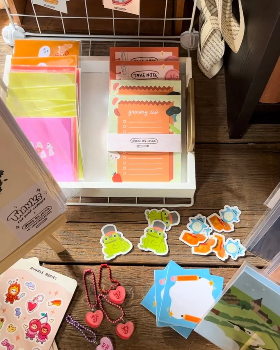 Colorful stationery and stickers on a wooden surface, including grocery list notes, frog stickers, blue note cards, and heart-shaped tags.