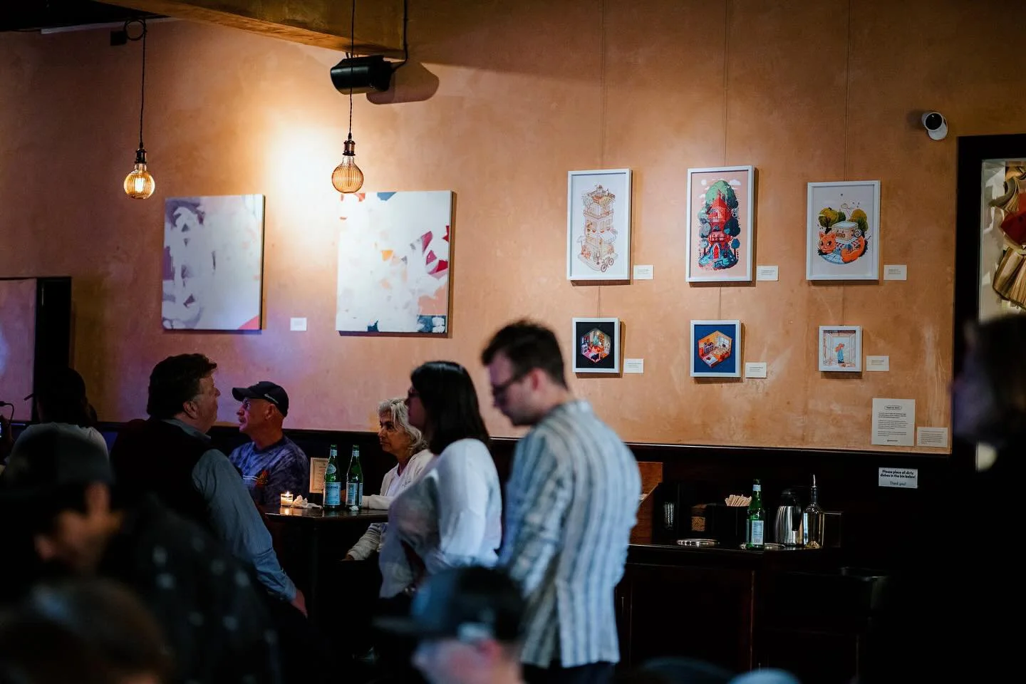 Interior of an art gallery or cafe with five people seated and engaging in conversation, and seven framed artworks on a peach-colored wall, illuminated by hanging light fixtures.