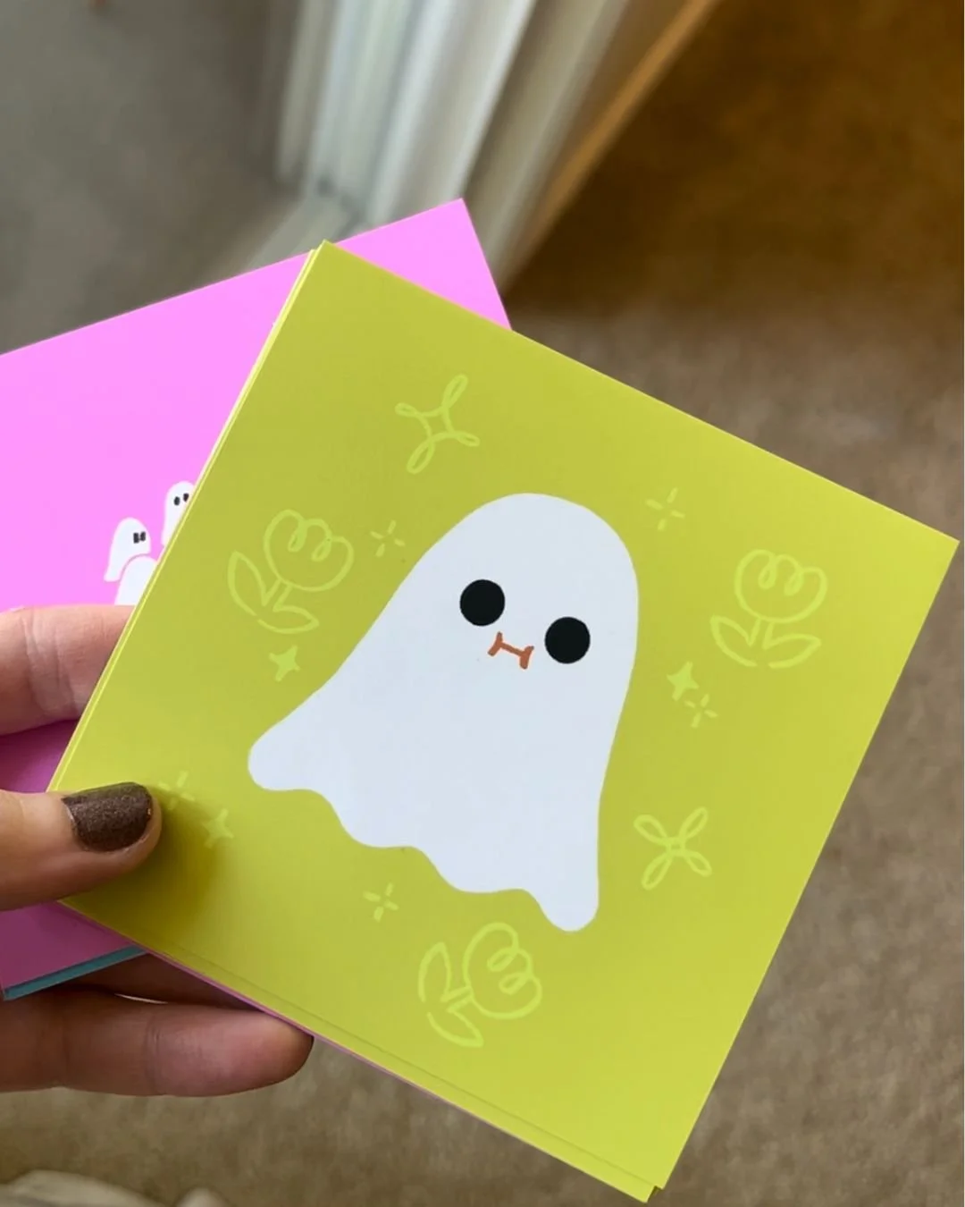 Hand holding a yellow note card with a cute ghost illustration, surrounded by flowers and sparkles, with a pink note card partially visible behind it.