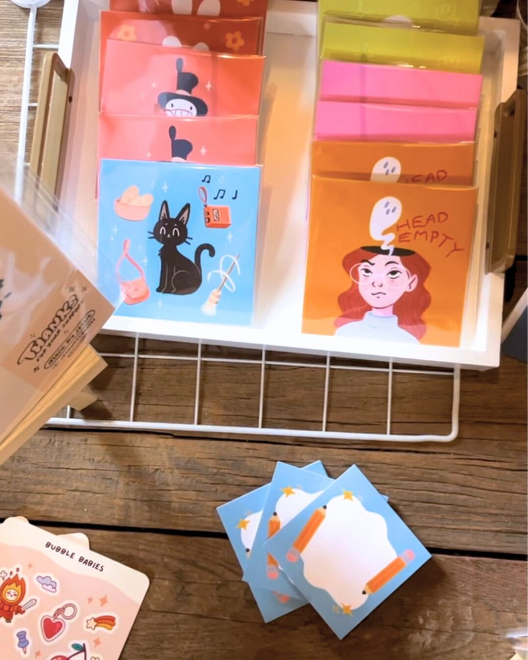 A display of colorful paper notebooks with various illustrations and messages, including a girl with a ghost saying "Head Empty," a black cat, and a girl with a ghost, alongside some decorative cards with illustrations of stars, a pencil, and a cloud