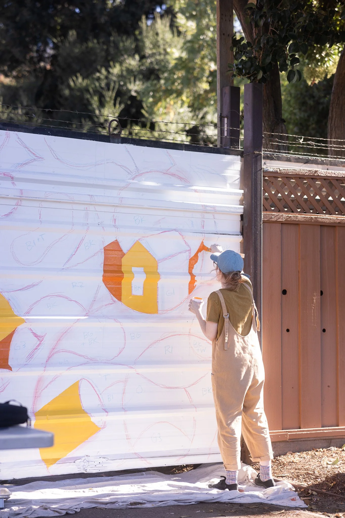 A person wearing beige overalls, a gray cap, and glasses is painting on a white corrugated metal wall outdoors. The wall has sketches with orange and yellow shapes, and the person is holding a small container of paint.