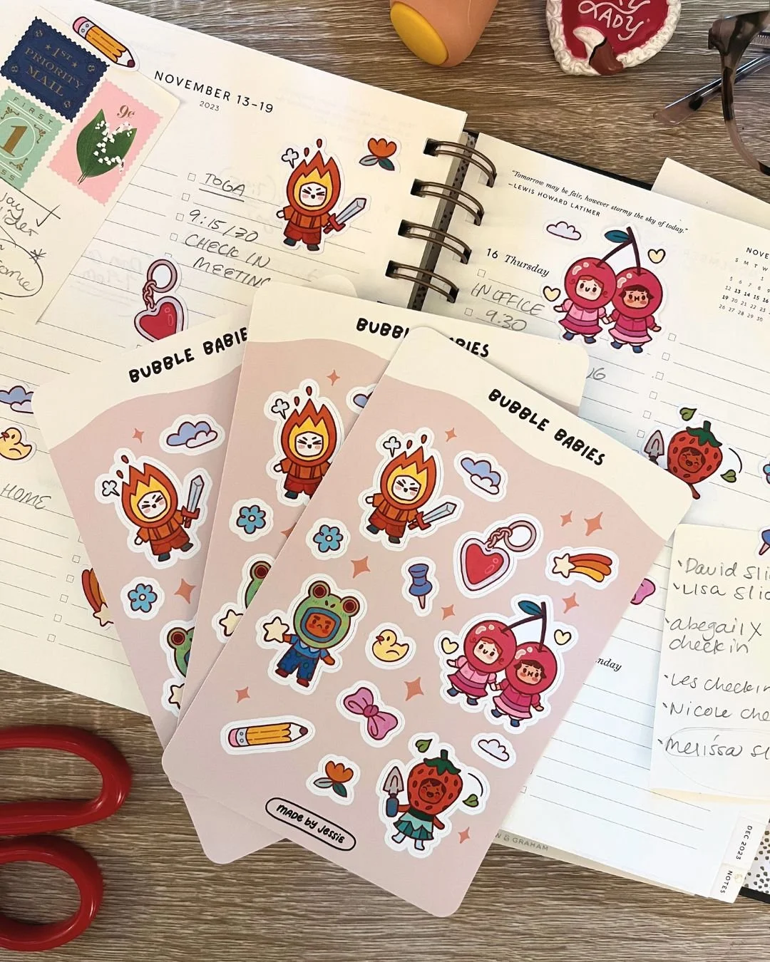 Three sheets of stickers with cartoon characters, including a person in a flame costume, a person in a bear costume, and a strawberry. The stickers also feature hearts, clouds, a pencil, a bow, stars, and a rainbow. The background includes a planner 