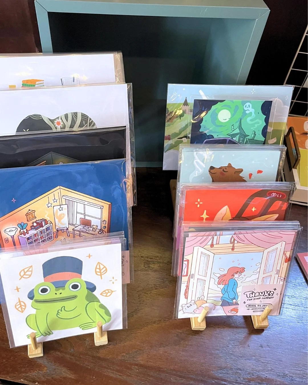 Display of colorful postcards with various illustrations, arranged on small wooden stands and cardholders, on a wooden table.
