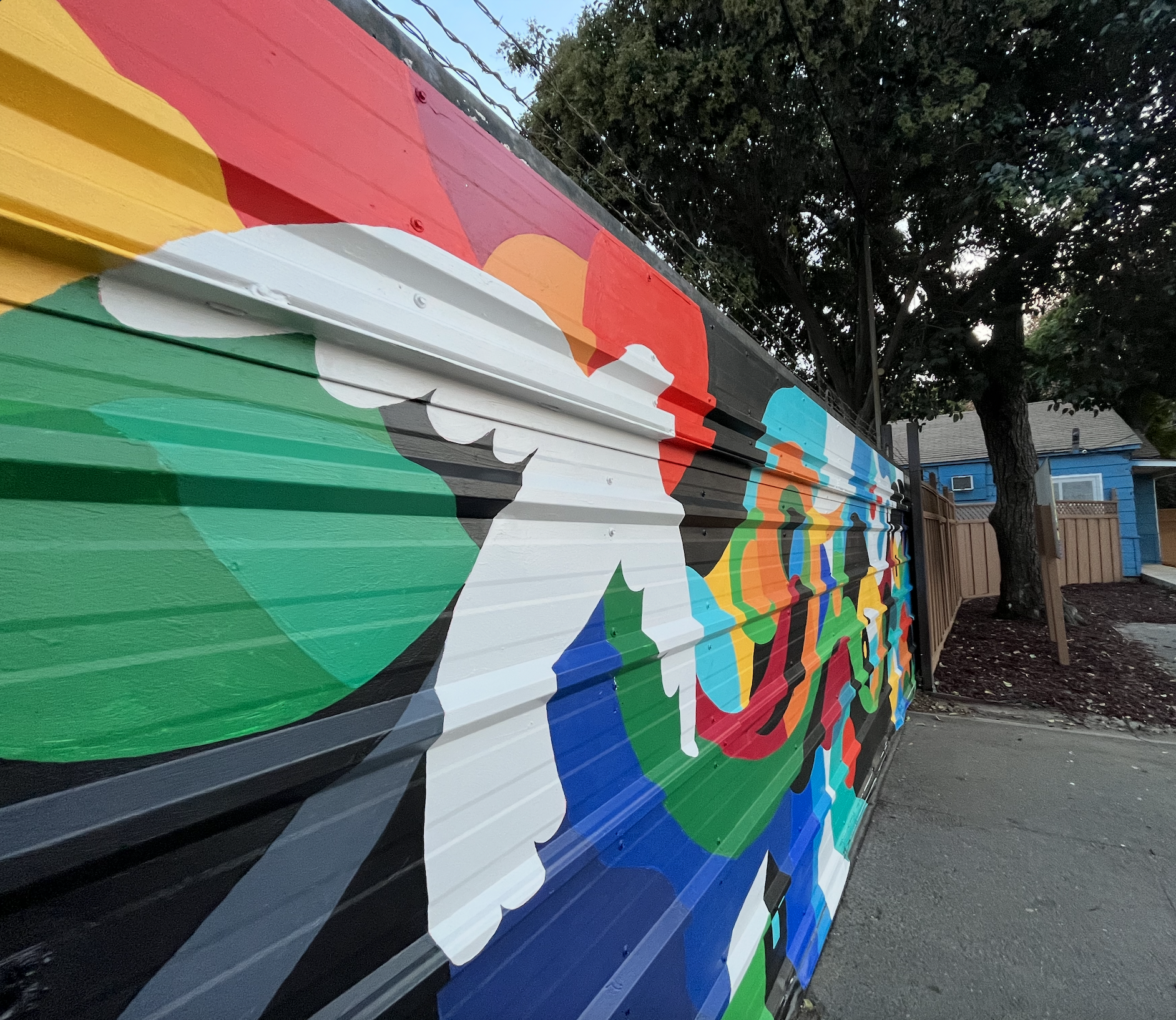 Colorful mural painted on a wooden fence featuring abstract shapes and figures, with trees and a house visible in the background.