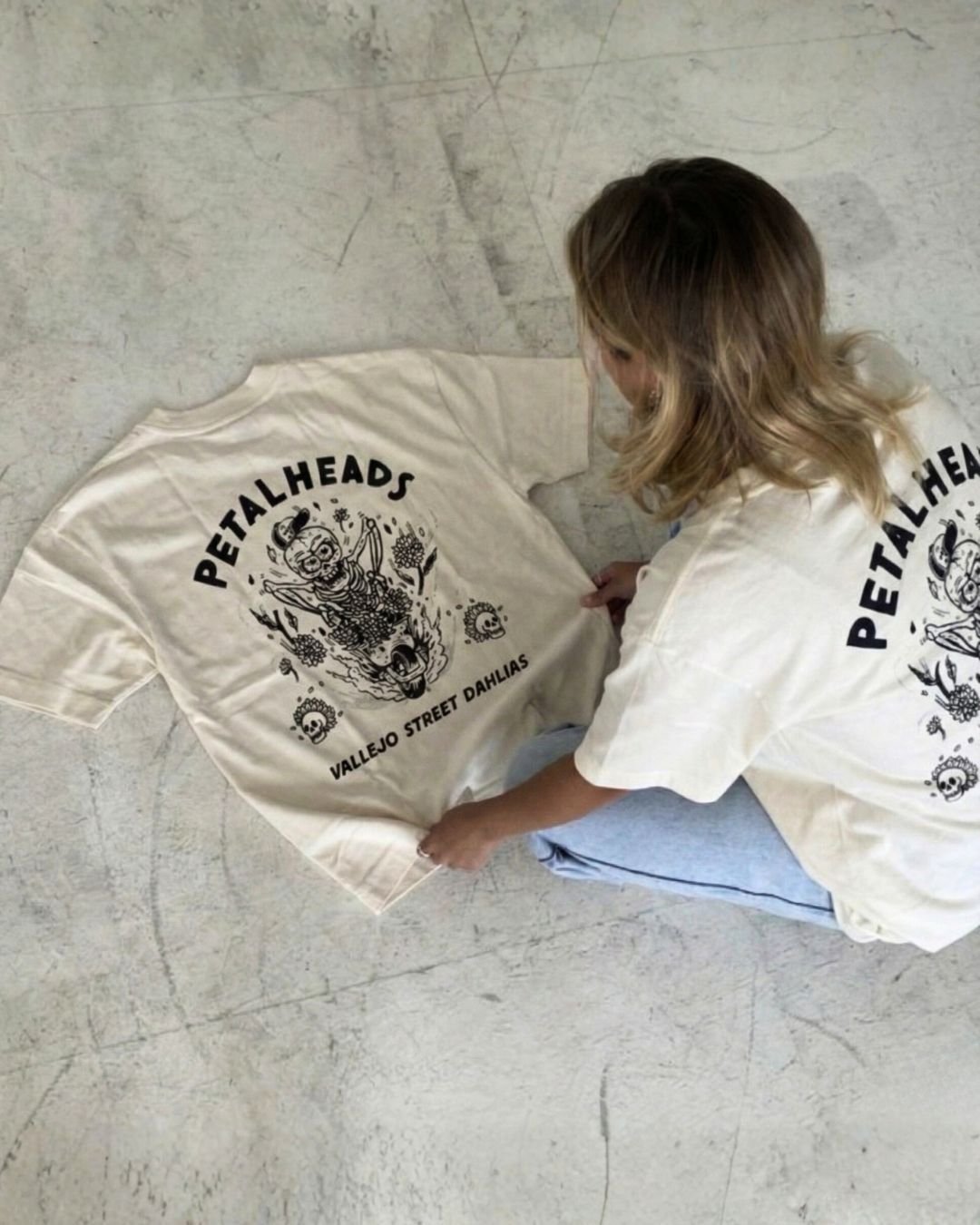A person folding a white T-shirt with a black graphic design featuring a skeleton and skulls, with text 'PETALHEADS' and 'VALLEJO STREET DAHLIAS' on it.