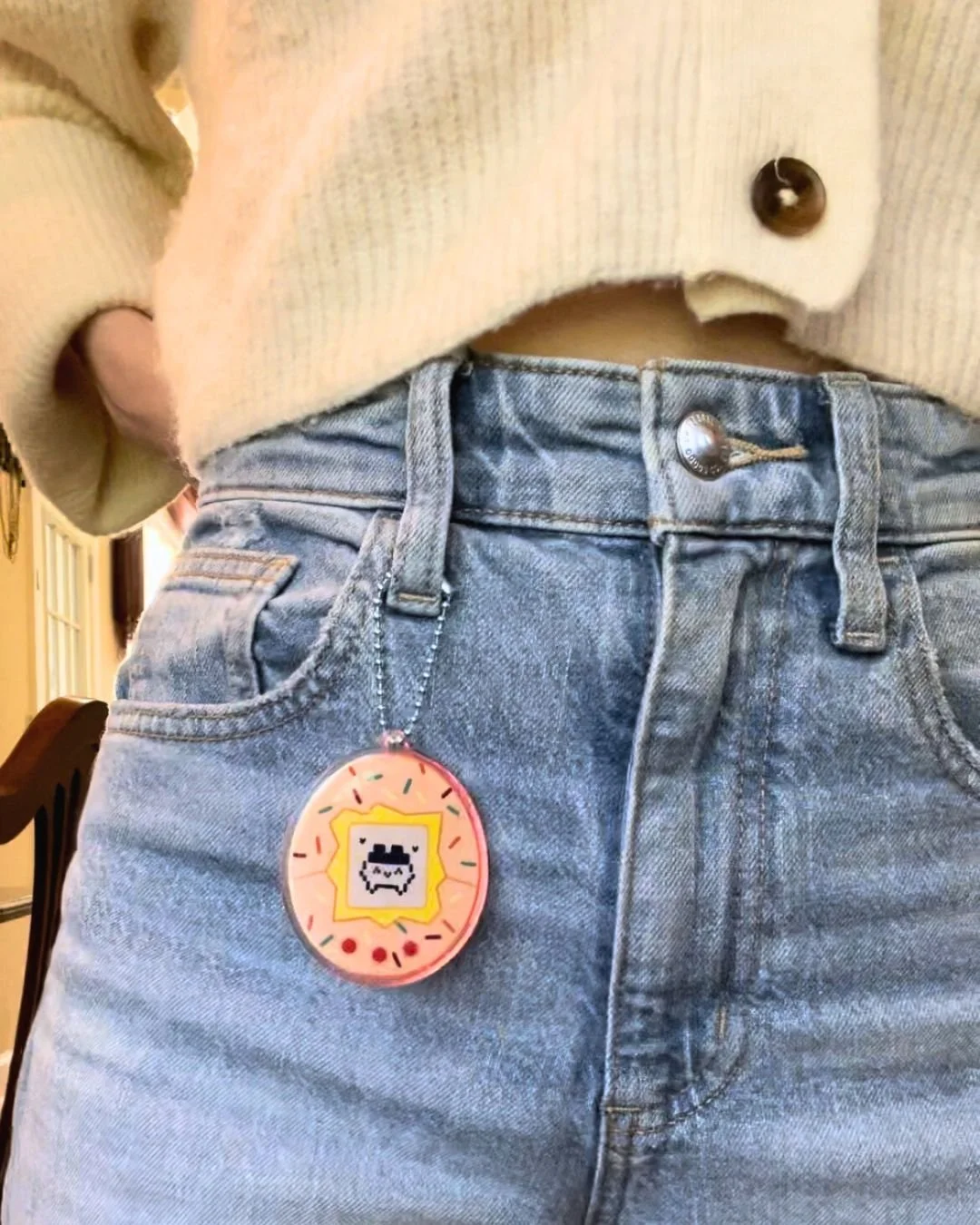 A close-up of a person wearing a beige cropped sweater and high-waisted blue jeans with a keychain attached to a belt loop. The keychain features a pixelated design of a smiling face inside a yellow frame, resembling a cartoon character.