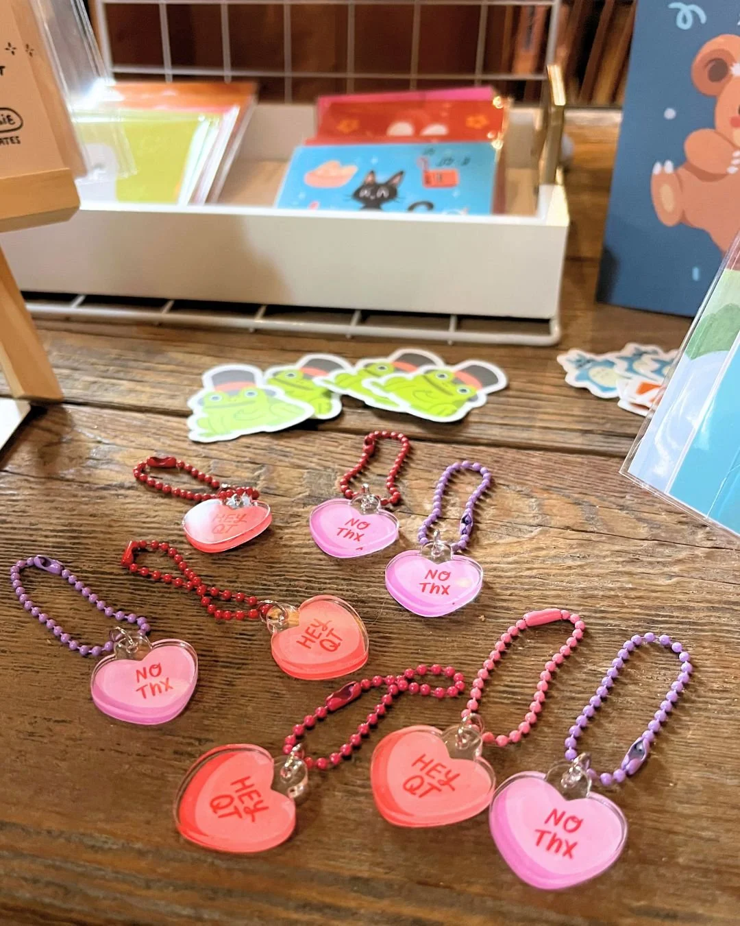 Display of colorful heart-shaped keychains with the words 'NO THX' and 'HEY QT' on a wooden surface, with assorted stickers and stationery in the background.