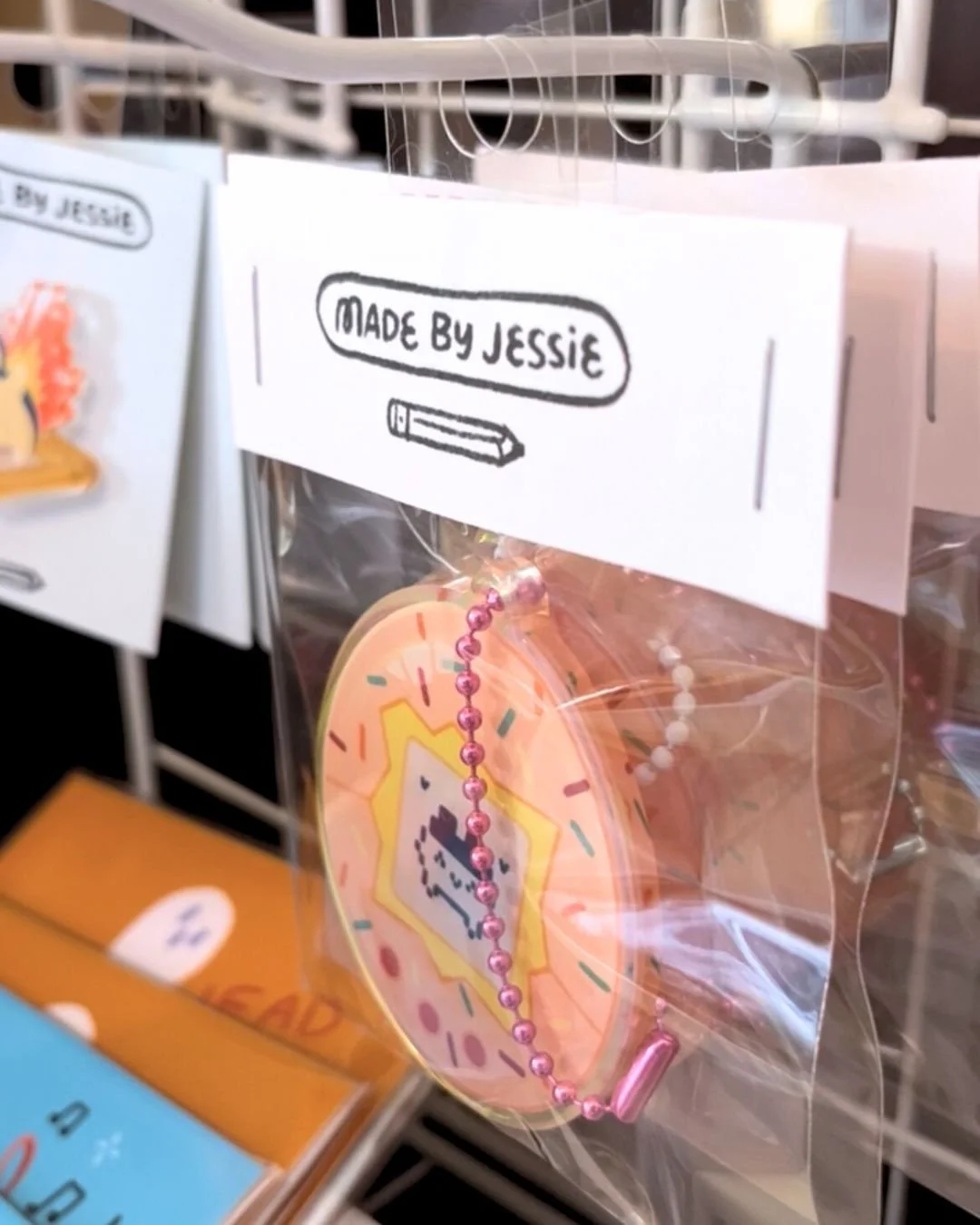 A clear plastic bag containing a round, peach-colored cookie with colorful sprinkles and a pink bead chain, labeled 'Made by Jessie' on a white tag with a black illustration of a crayon.