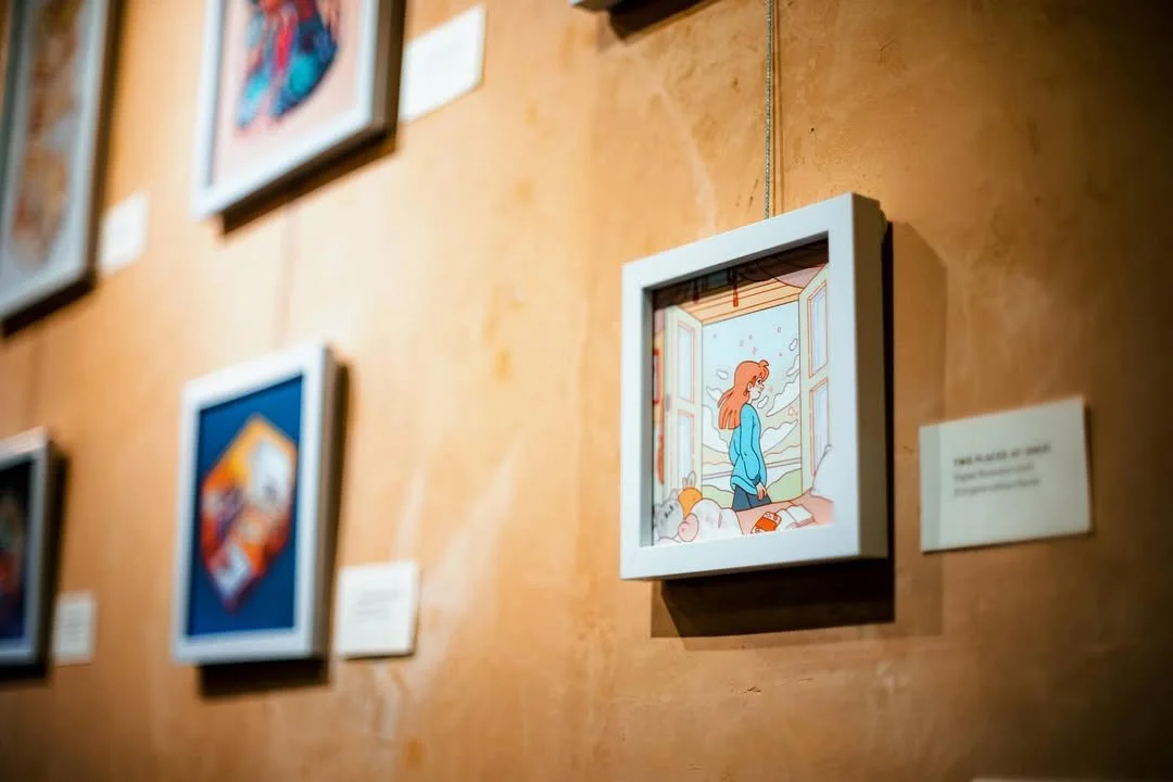 A framed digital illustration of a woman with long red hair standing by an open window, looking outside on a cloudy day. The artwork is displayed on a gallery wall with other framed pieces.