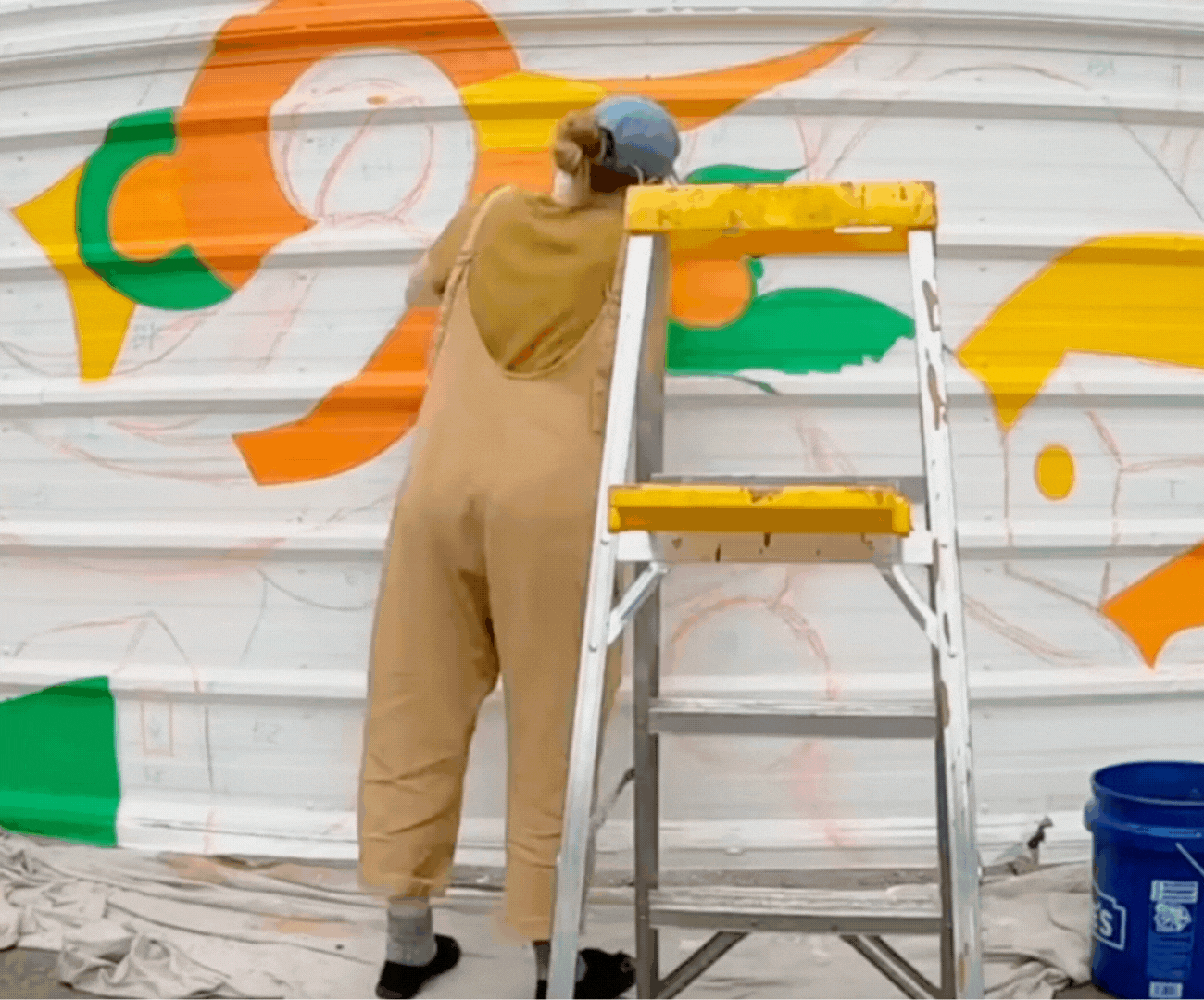 A person painting a colorful mural on a wall using a step ladder, with the mural featuring bright orange, green, and yellow shapes.