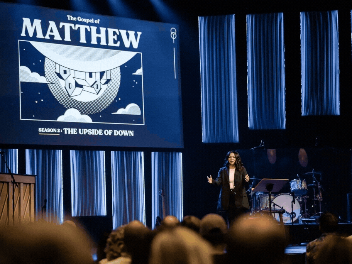 A woman speaking on stage in front of a large screen displaying promotional artwork for 'The Gospel of Matthew,' season 2 titled 'The Upside of Down,' with blue curtains in the background and an audience in the foreground.