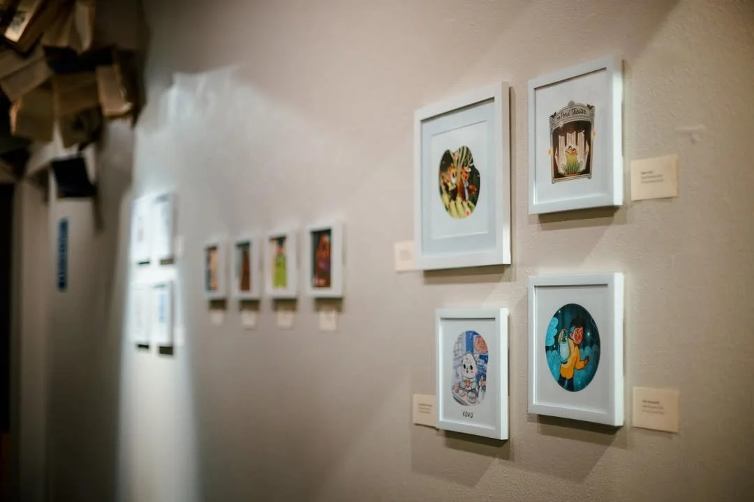 An art gallery wall displaying framed artworks, including illustrations of animals, fantasy scenes, and characters, arranged in a mostly horizontal line with small informational placards next to each piece.