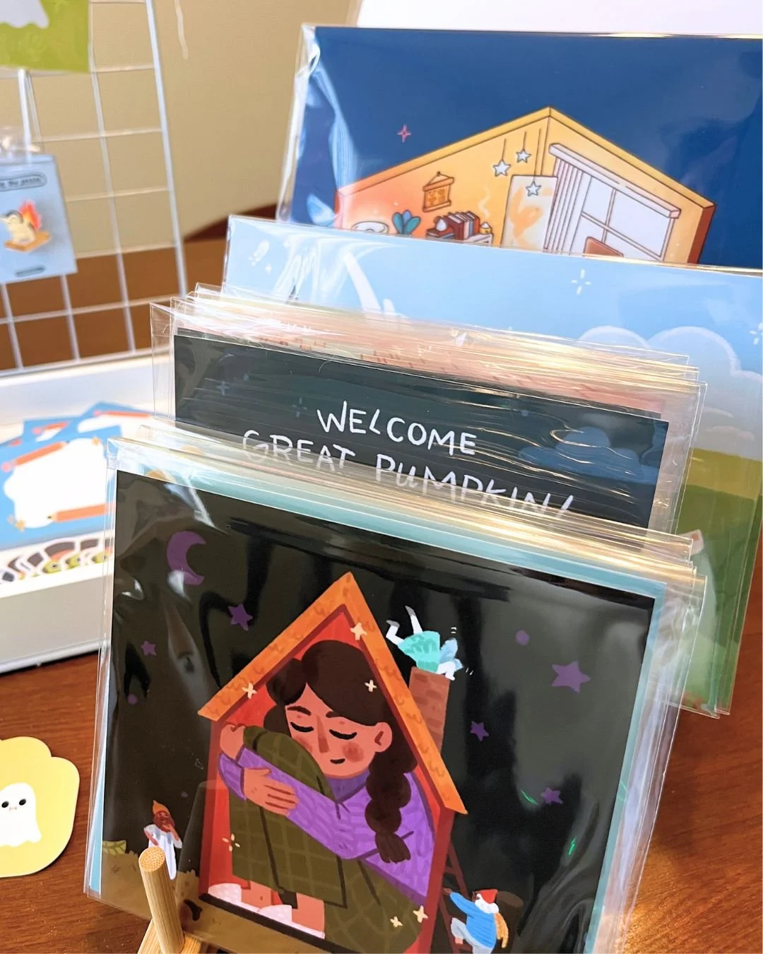 A collection of colorful greeting cards in clear plastic sleeves displayed on a wooden surface. One card features a girl hugging a cat inside a cozy house with stars around, and other cards have various designs including welcoming messages.