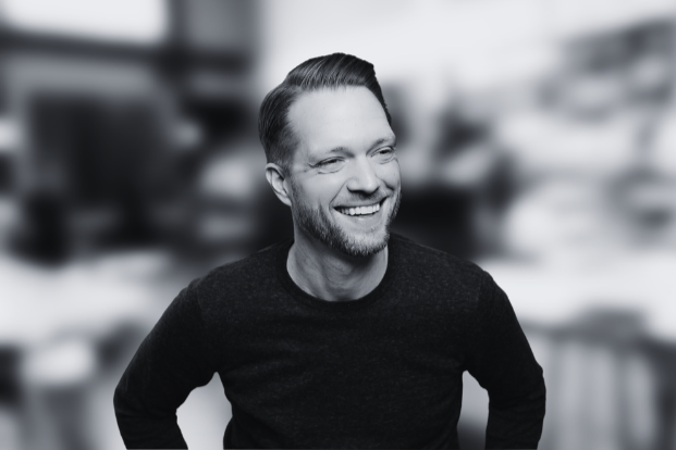 A smiling man with a beard and slicked-back hair in a black sweater, standing in a blurred outdoor setting.