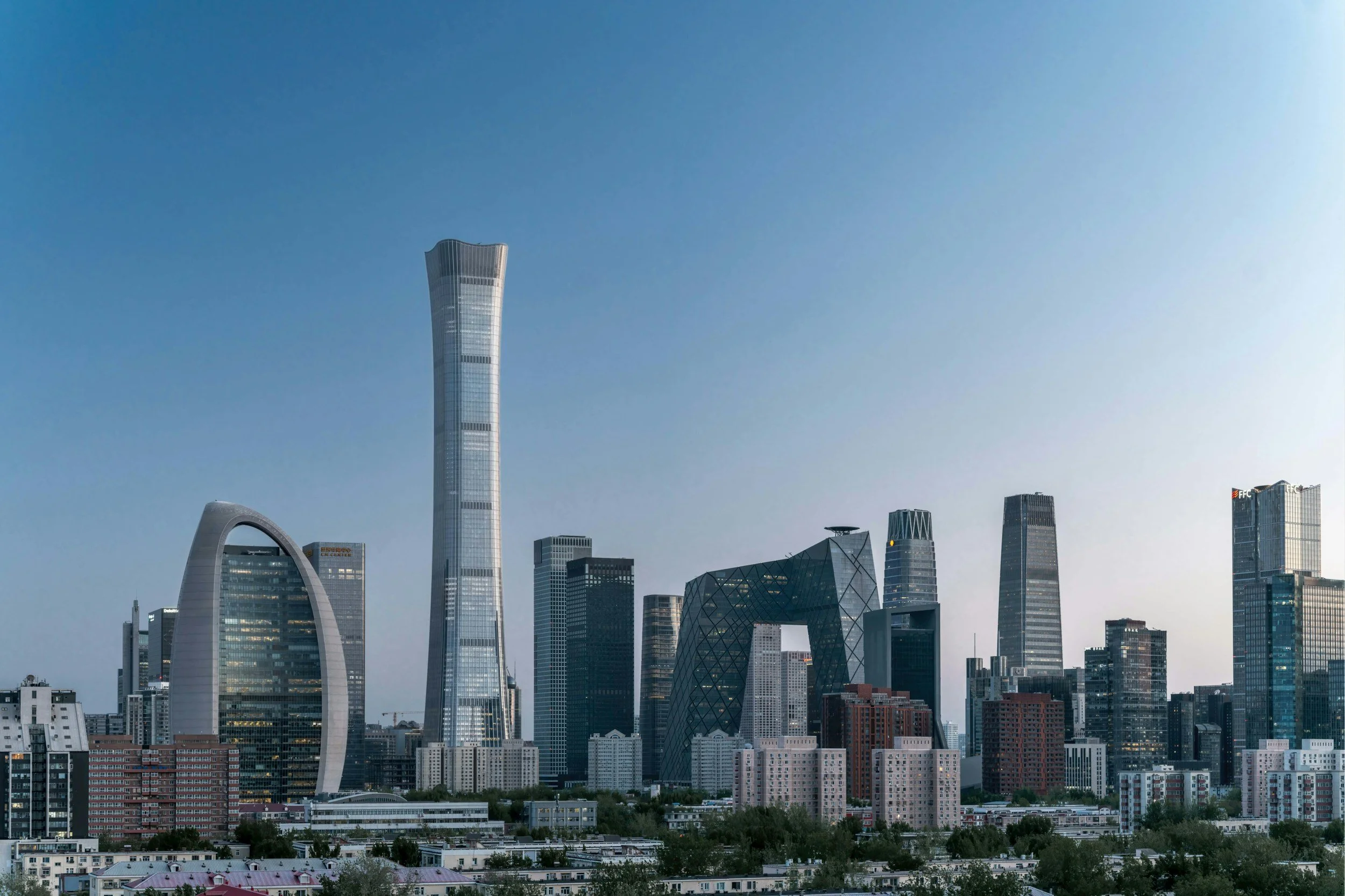 A city skyline with tall modern skyscrapers, including the China Zun Tower, Beijing's tallest building, against a clear blue sky.