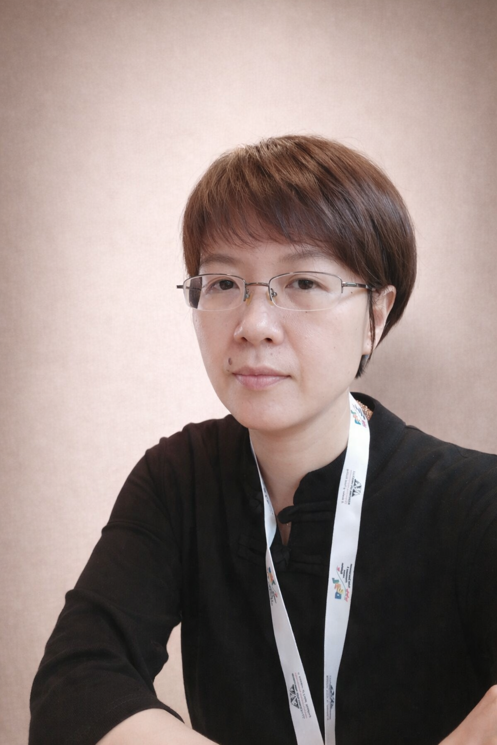 A woman with short brown hair wearing glasses and a black shirt, taking a selfie in front of a plain beige wall.