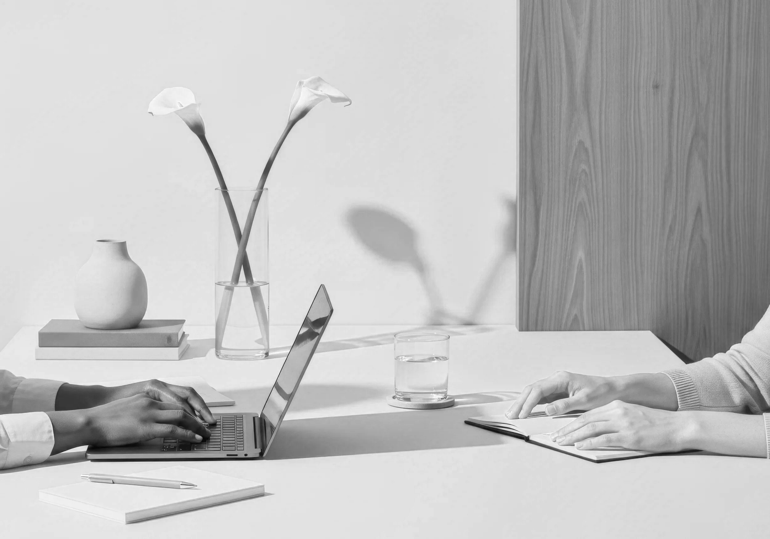 Two people are sitting across from each other at a table. One person is typing on a laptop, and the other is holding a notebook. There is a glass of water, a pen, a vase with two flowers, and books on the table. The background features a wooden wall panel and a white wall.