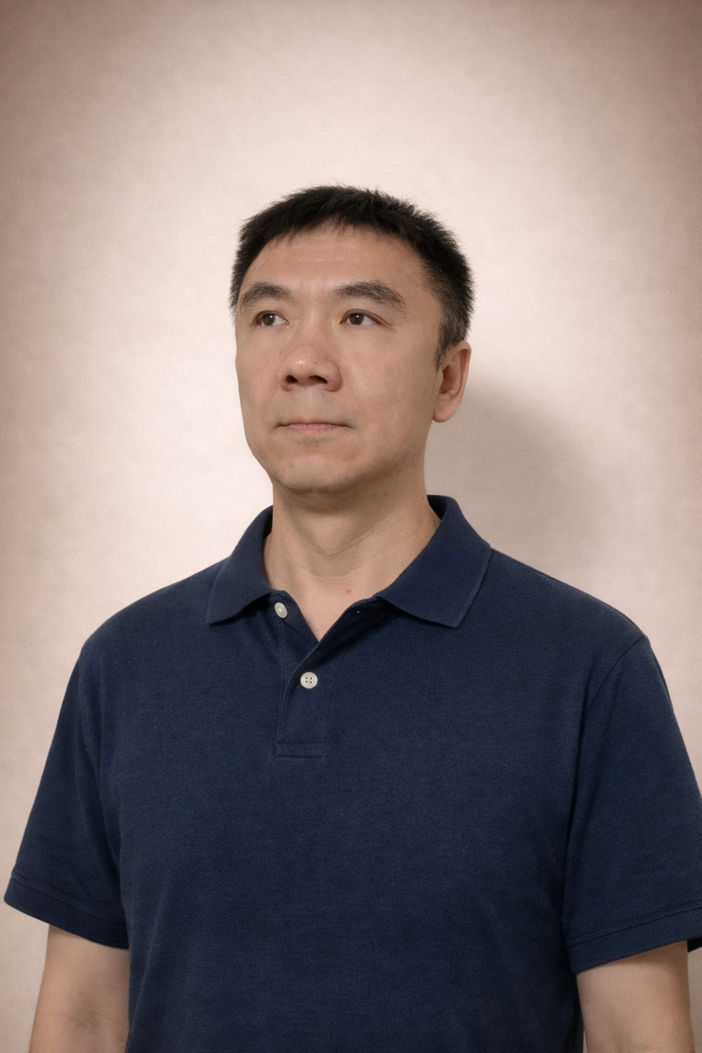A man with short black hair, wearing a navy blue polo shirt, looking slightly to the right against a plain light-colored background.