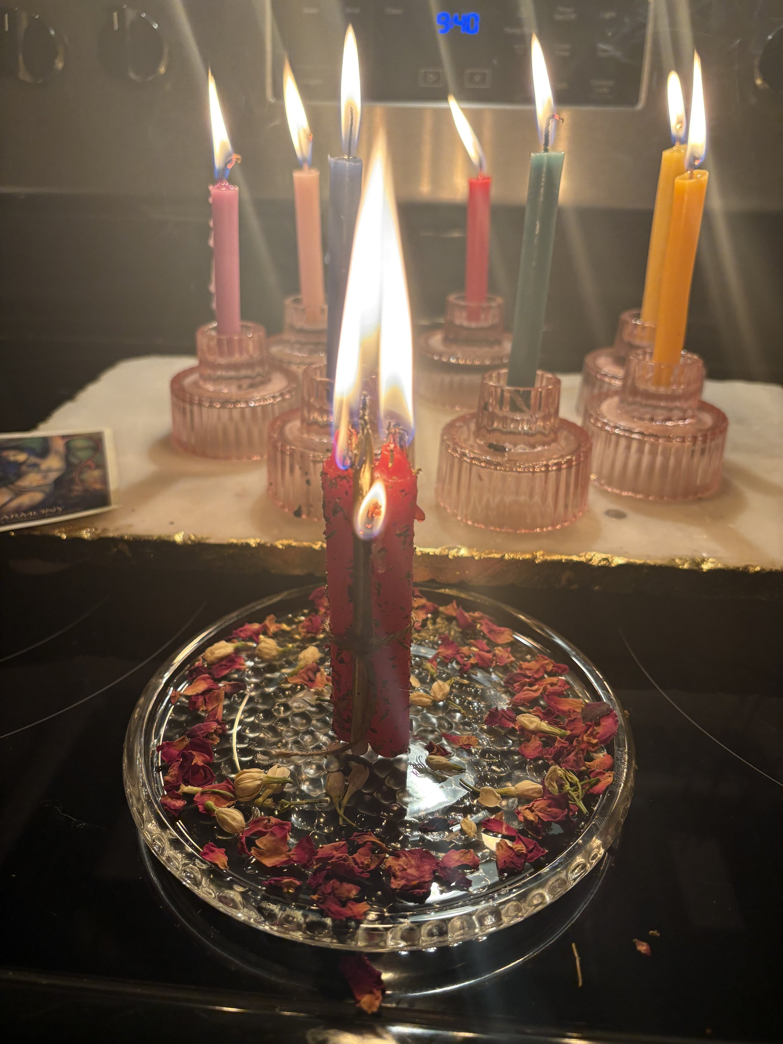 A birthday cake with red and black candles and flower petals on top, with multiple lit candles. There are additional unlit candles in the background.