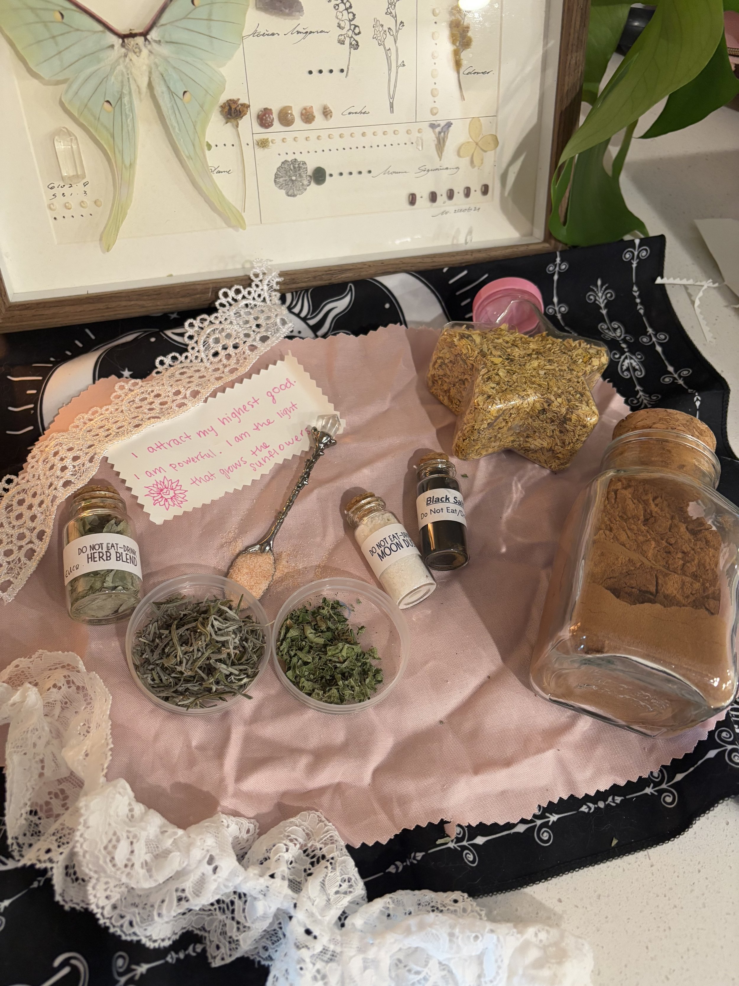 A collection of dried herbs, spices, and crystals arranged on a pink cloth, with a handwritten note that says, 'I attract my highest good. I am powerful. I am the light that gives the sunflowers their sunshine.' In the background, a framed botanical and jewelry display and a green plant are visible.