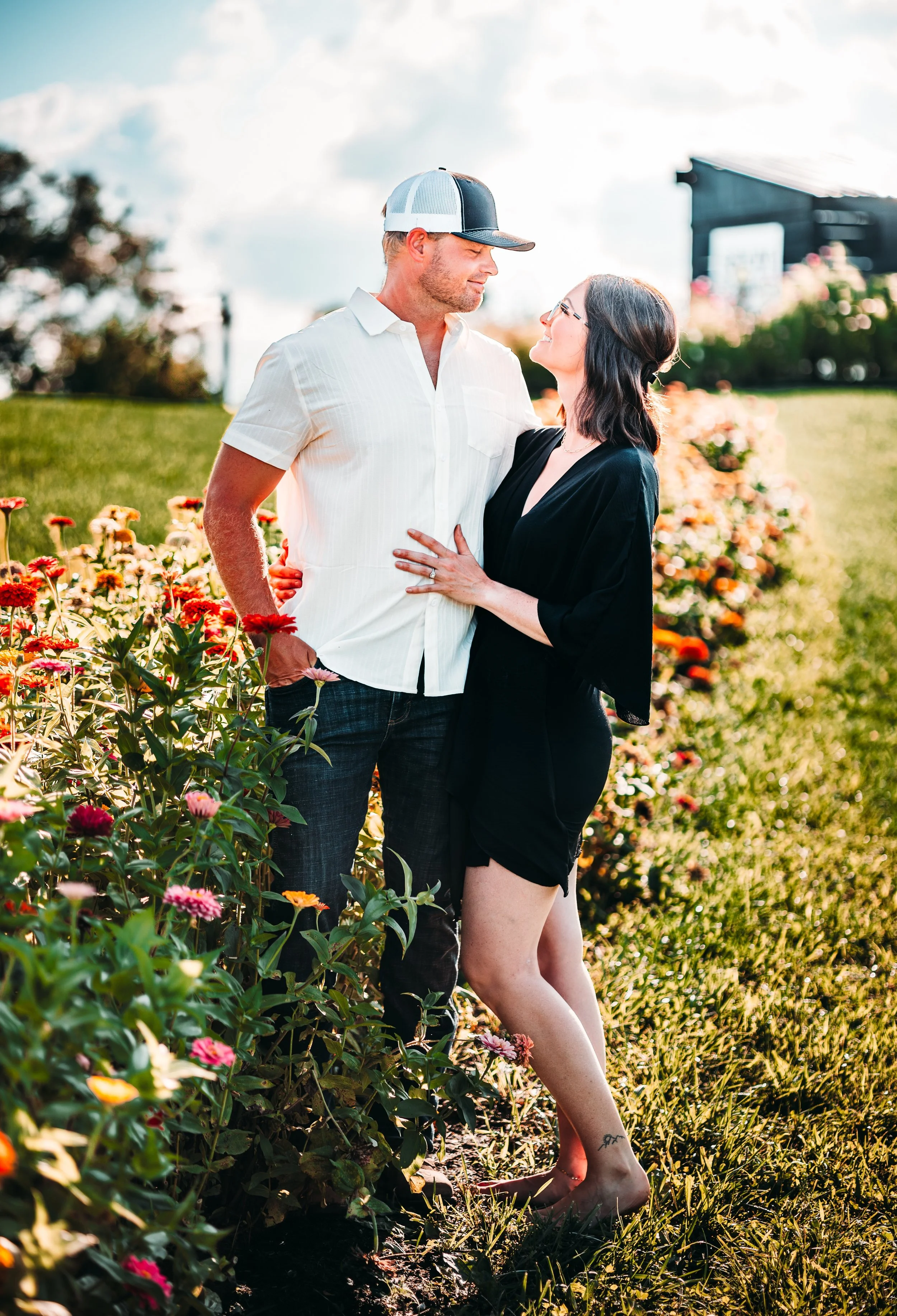 A man and woman are standing close together in a garden with colorful flowers. The man is wearing a white short-sleeve shirt, jeans, and a baseball cap, while the woman is in a black dress. They are looking at each other affectionately, with the woman touching the man's chest. The sky is partly cloudy, and the scene is brightly lit.