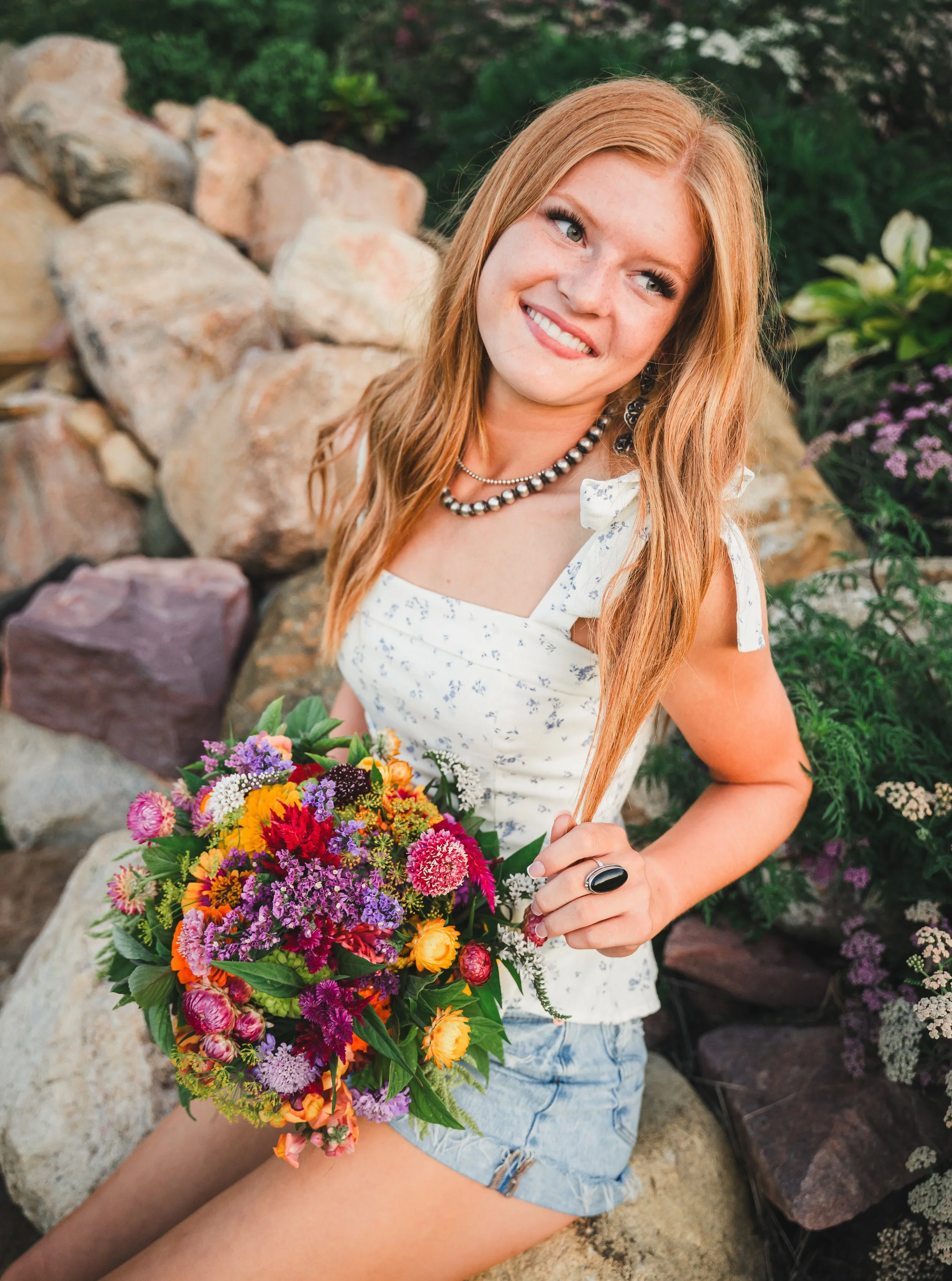 A young woman with long red hair sitting outdoors on rocks, holding a colorful bouquet of flowers, wearing a white sleeveless top with a small floral pattern, denim shorts, a pearl necklace, and a large black ring.