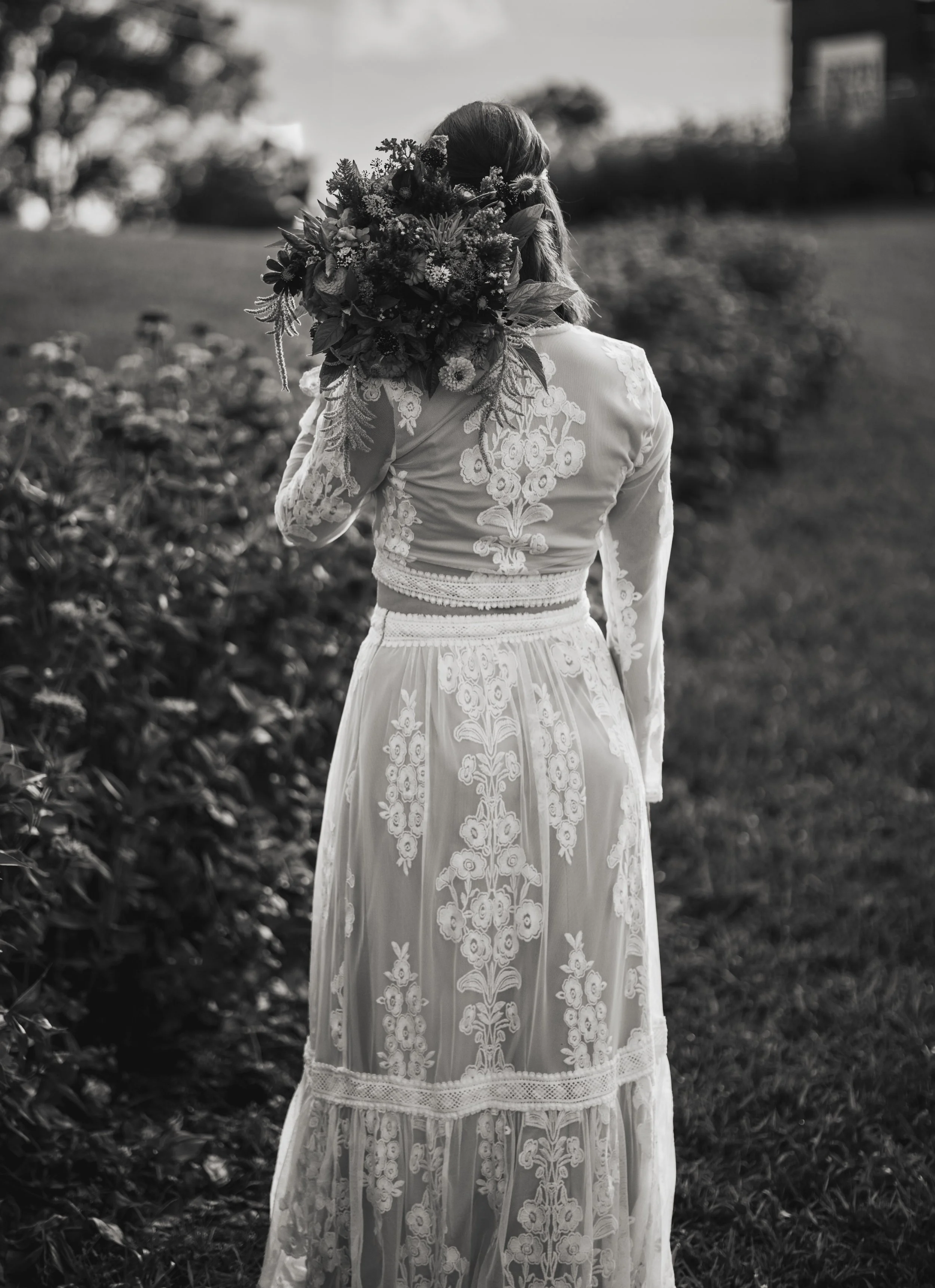 A woman in a white, embroidered dress carrying a large bouquet of flowers outdoors in a garden or park setting.