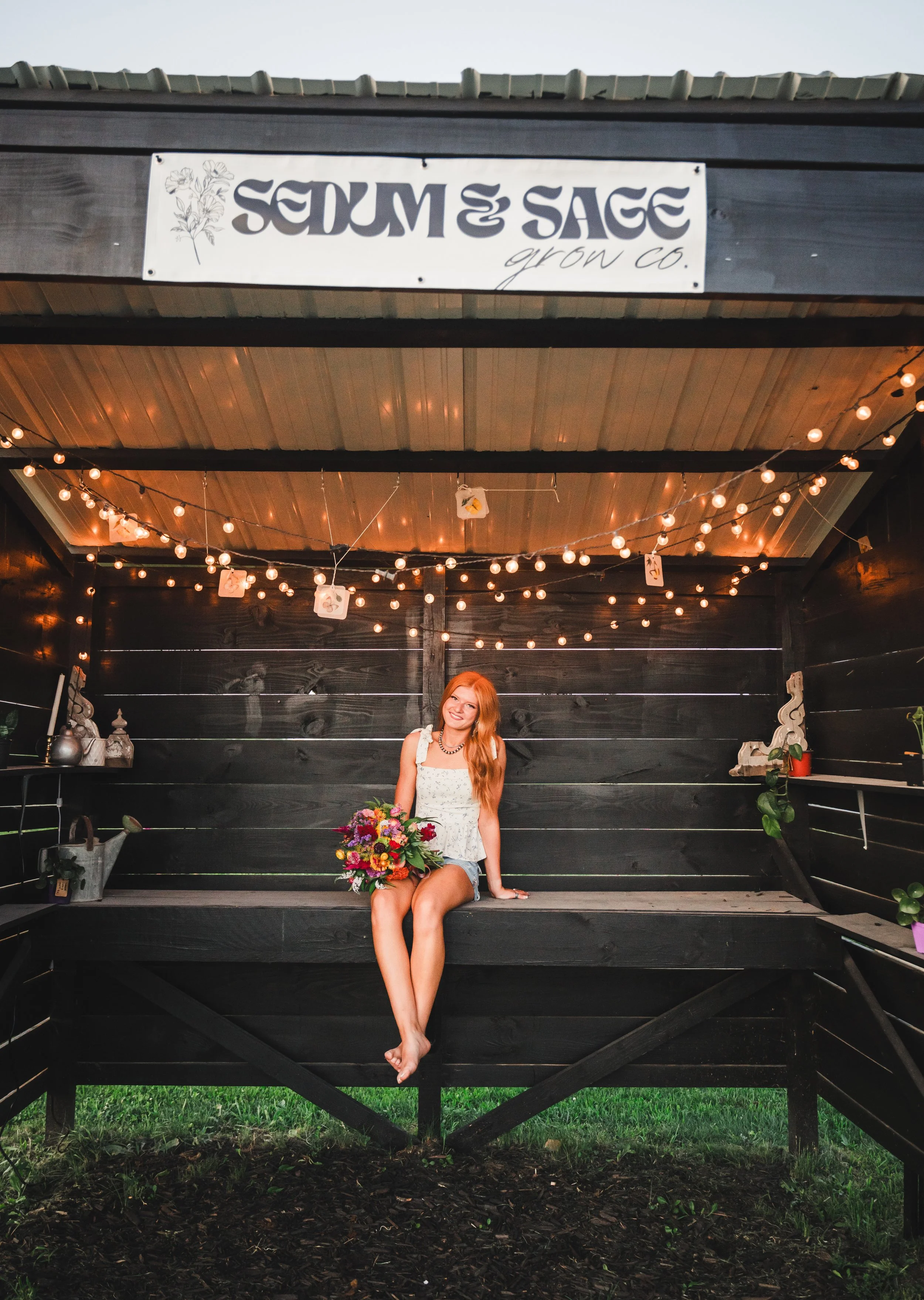 A young woman with red hair sitting inside a black wooden structure, holding a colorful bouquet of flowers, under string lights, with a sign above that reads 'Sedum & Sage grow co.'