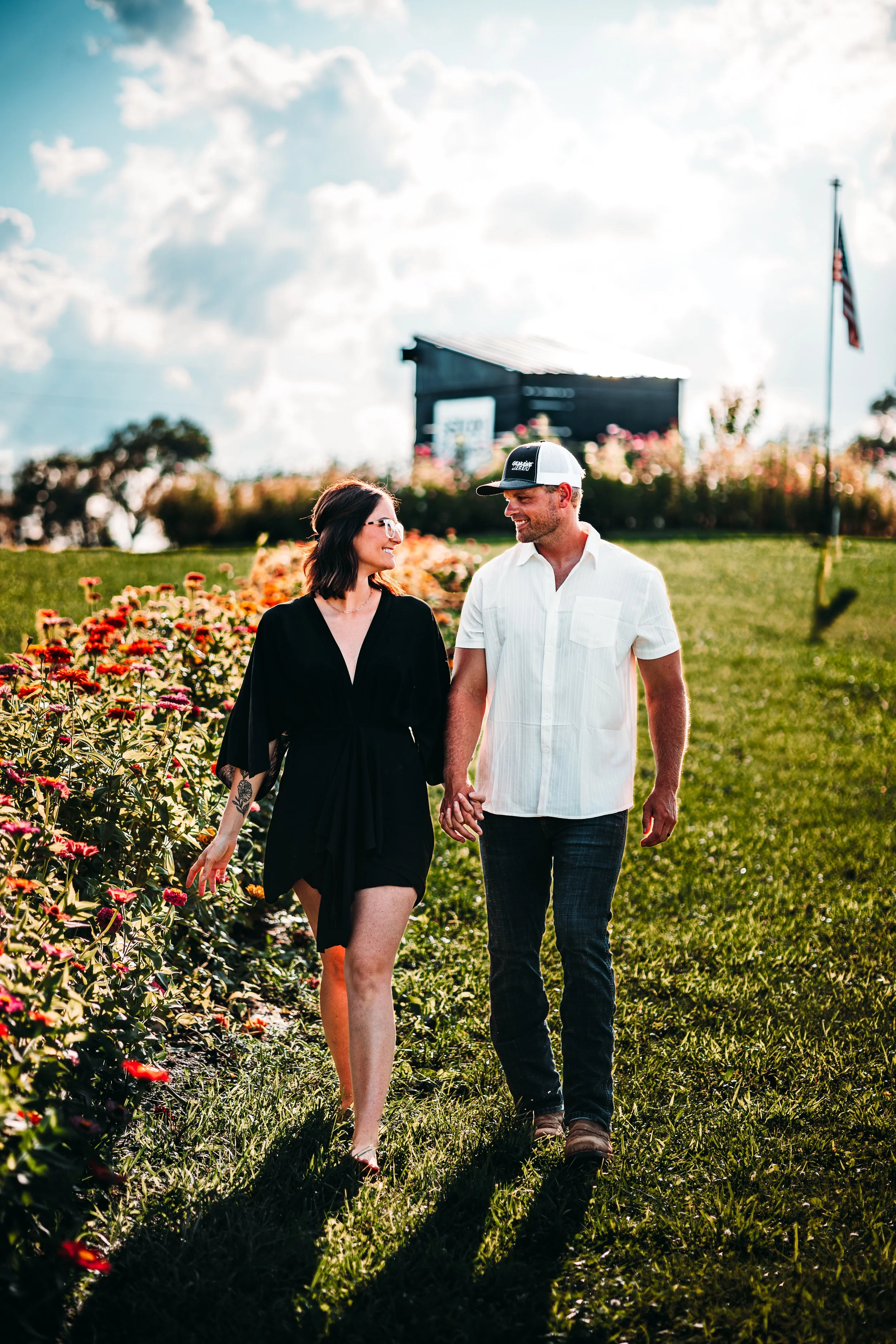 A couple walking hand in hand in a flower garden on a sunny day, with a barn and an American flag in the background.