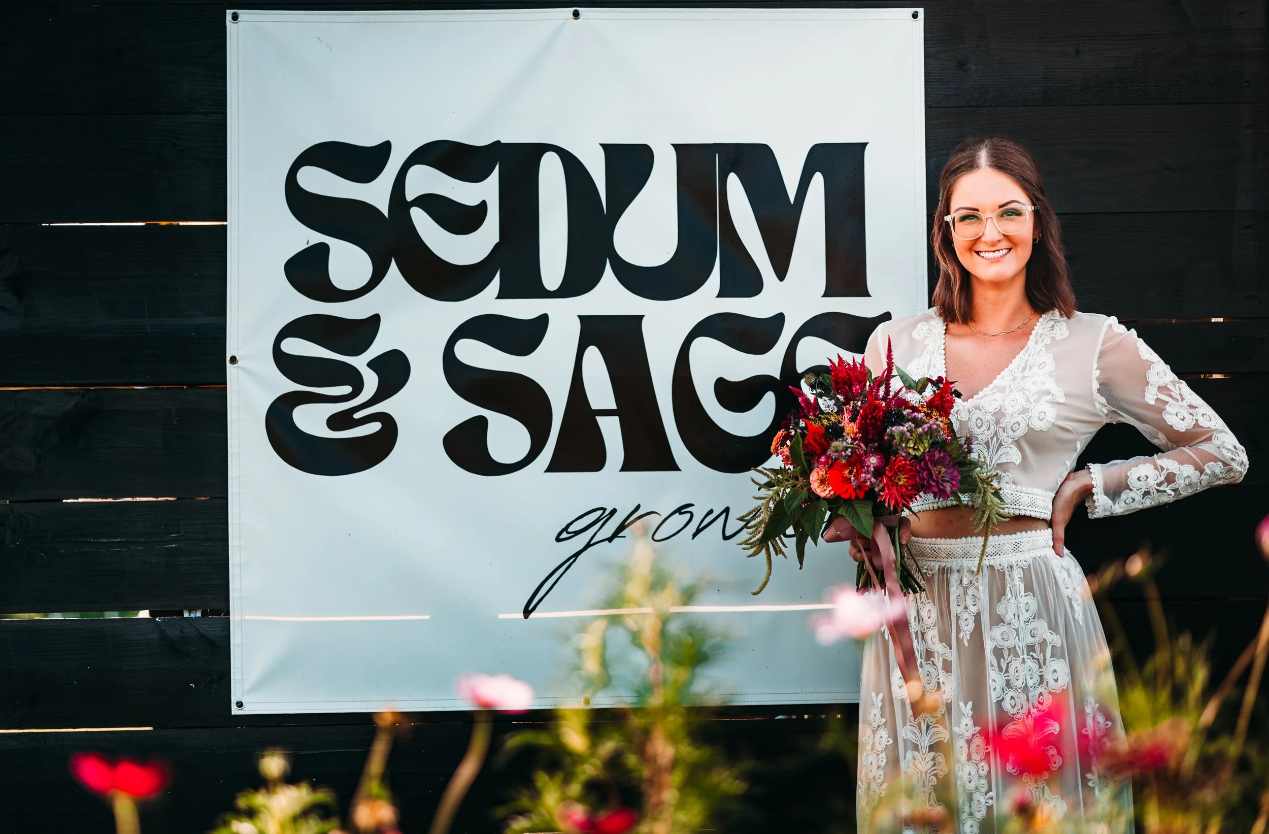 Woman smiling and holding a bouquet of flowers at an outdoor event, standing next to a sign that says "SUM & SACKGROWNS" on a black wooden wall.