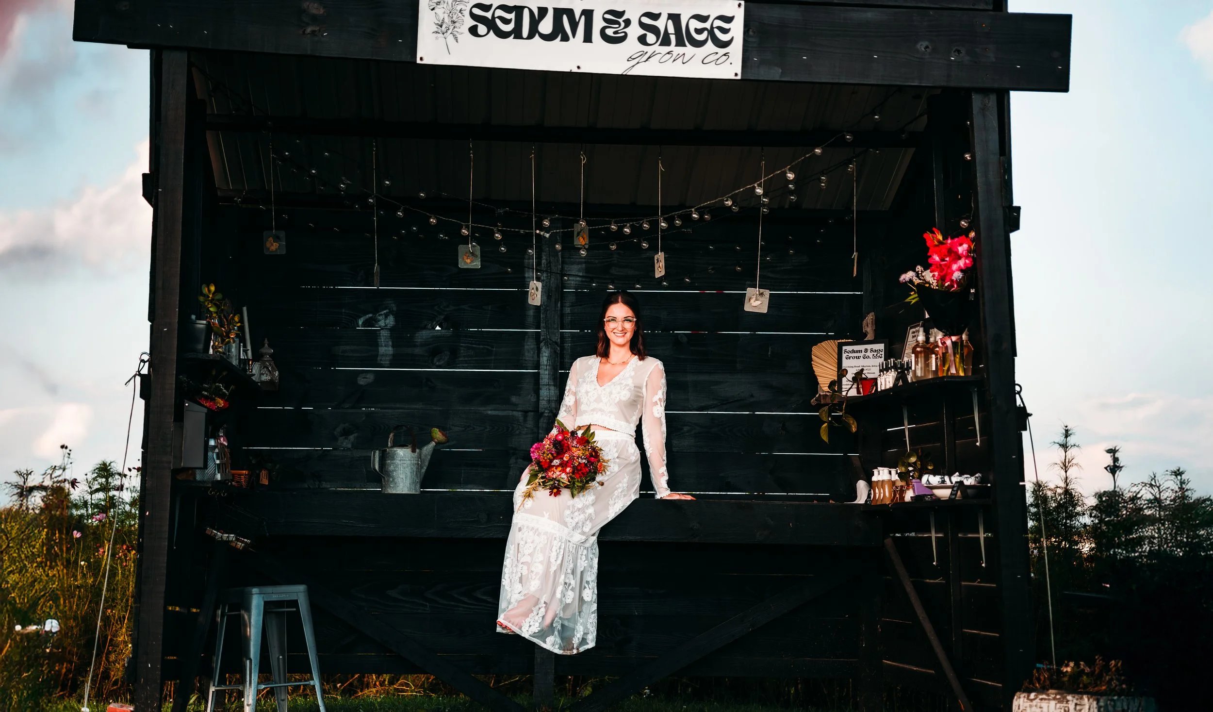A woman in a white lace dress sitting on the edge of a black wooden food stand with a bouquet of flowers, at sunset with a cloudy sky and trees in the background, under a sign that reads 'Sedum & Sage Grow Co.'.