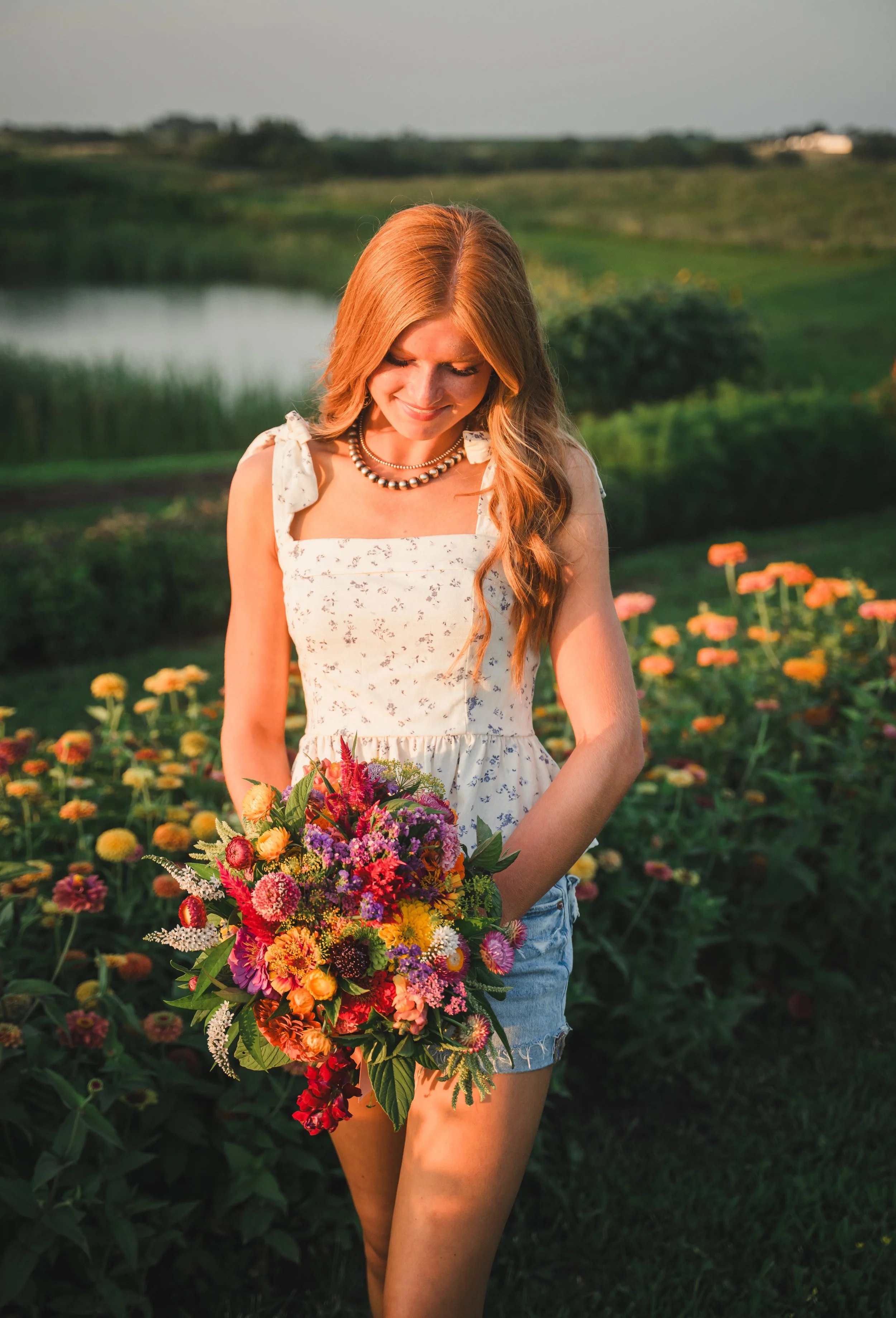 A woman with long, wavy red hair smiling and holding a colorful bouquet of flowers while standing in a field of flowers near a pond during sunset.
